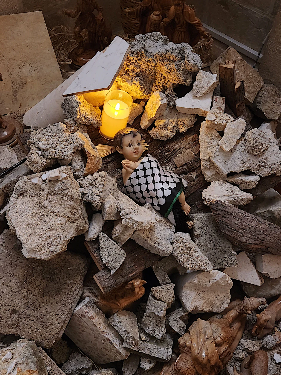 Installation statt Weihnachtsbaum: Ein Haufen Steine, darum herum Holzfiguren, in der Mitte liegt eine Puppe eingehüllt in einen schwarz-weißen Palästinenserschal. Angehörige der evangelisch-lutherischen Weihnachtskirche wollen damit an die unter Trümmern verschütteten Kinder im Gazastreifen erinnern.