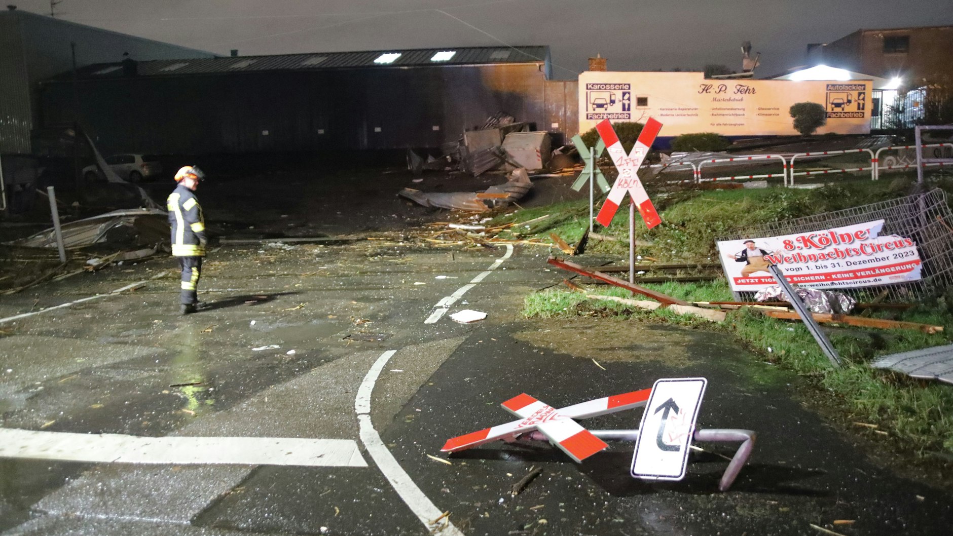 Ein Feuerwehrmann betrachtet die Trümmer und Schäden an einem Bahnübergang in Köln. Zahlreiche Schilder sind umgeknickt und Dächer abgedeckt. 