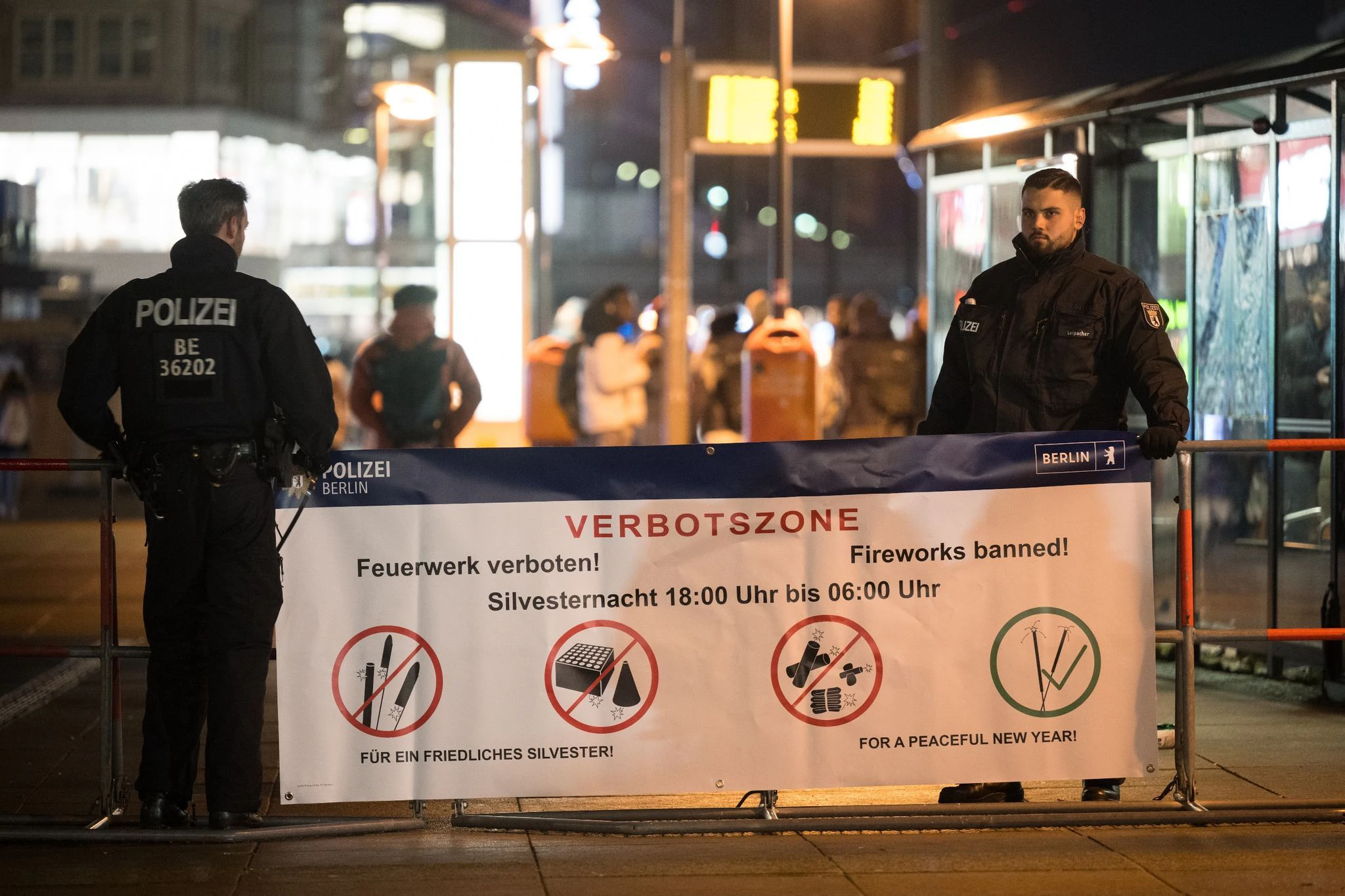 Polizeibeamte stehen an einem Plakat, das auf die Böllerverbotszone am Berliner Alexanderplatz hinweist.  