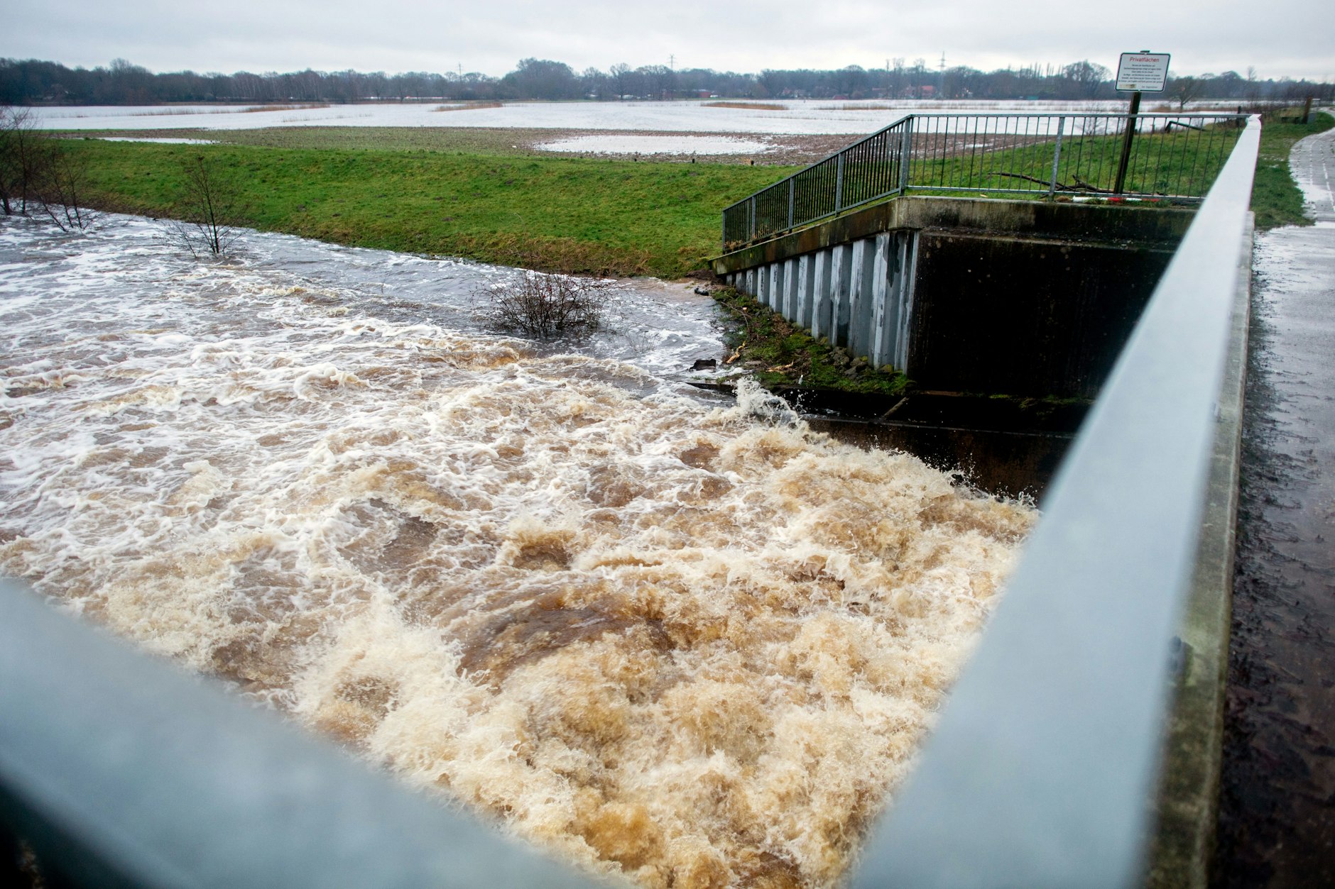 Wasser der Hunte strömt an einem Wehr in den Osternburger Kanal, während die Wiesen der Huntemarsch im Hintergrund überschwemmt sind.