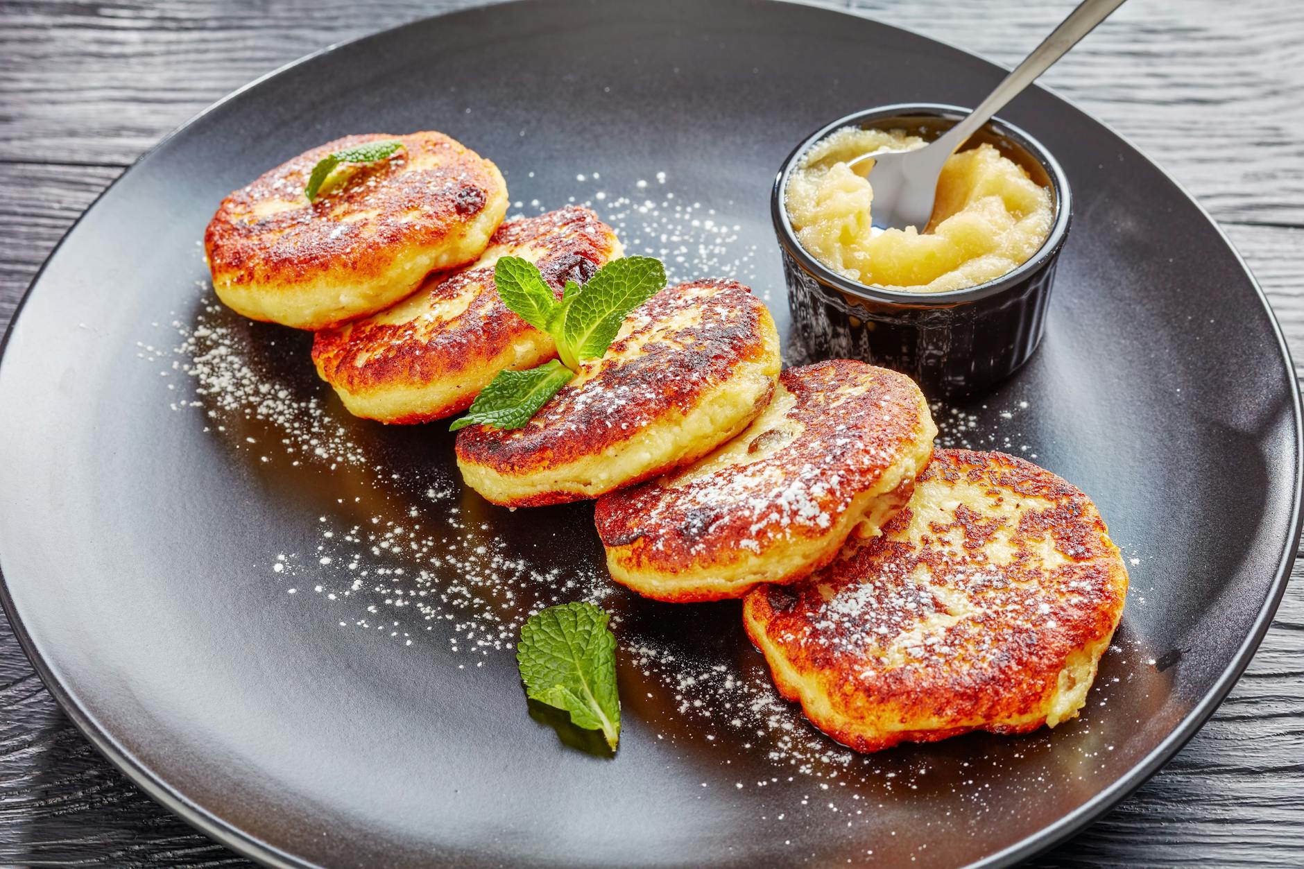Frisch gebratene Quarkkeulchen auf einem Teller, serviert mit Apfelmus und bestäubt mit Puderzucker.