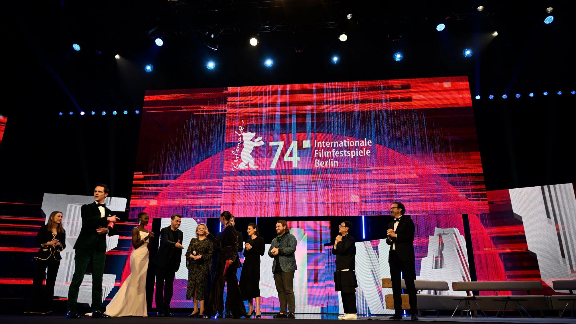 Die Berlinale-Leitung Mariette Rissenbeek und Carlo Chatrian (beide jeweils außen), die Berlinale-Jury sowie die Moderatoren Hadnet Tesfai und Jo Schück (2. u. 3. v. l.) stehen am Eröffnungsabend der Berlinale auf der Bühne.