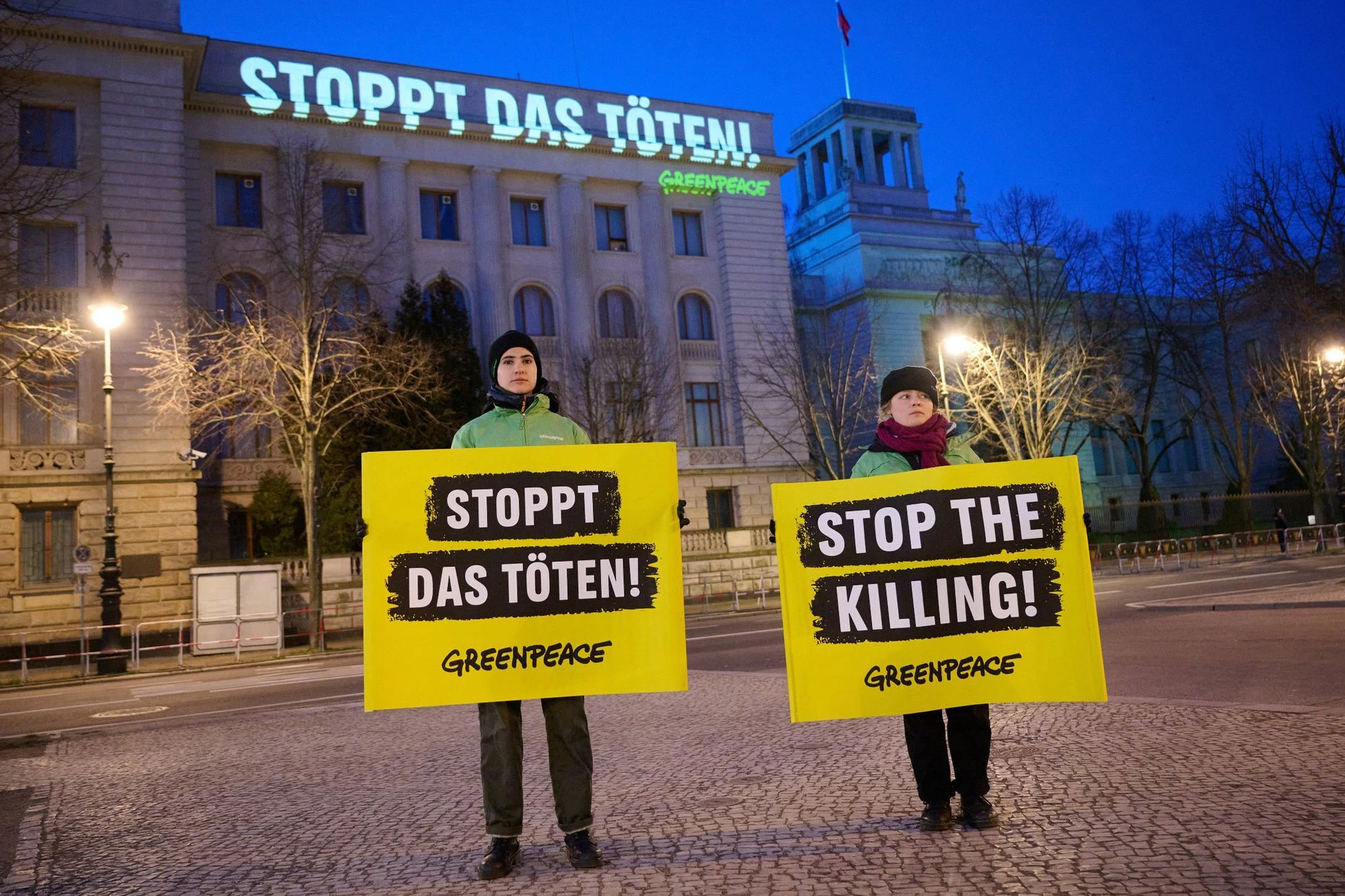 Greenpeace projiziert den Satz „Stoppt das Töten!“ an die Fassade der russischen Botschaft in Berlin. Aktivistinnen wiederholen davor die Aufforderung mit Plakaten.&nbsp;