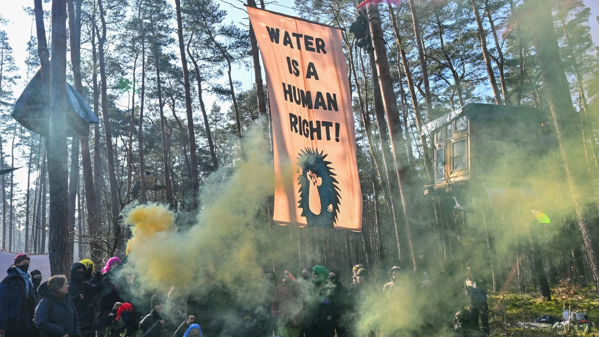 Aktivisten der Initiative „Tesla stoppen“ stehen mit Rauchfackeln vor einem Transparent mit der Aufschrift „Water is a human right!“ in einem Kiefernwald nahe dem Werk der Tesla-Gigafactory Berlin-Brandenburg.&nbsp;