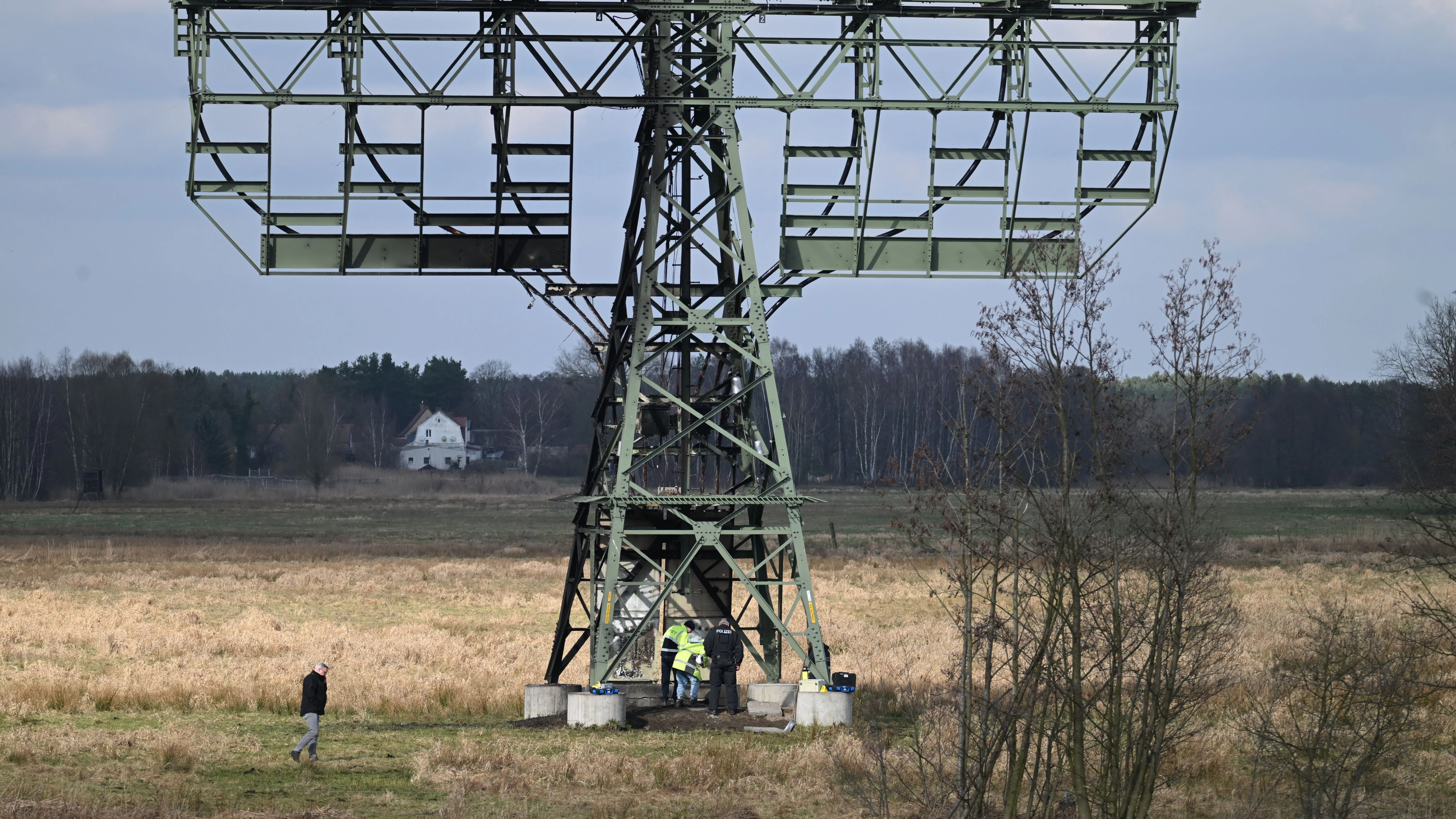 Die Polizei ermittelt an einem beschädigten Strommast auf einem Feld. In der Tesla-Autofabrik in Grünheide steht die Produktion wegen des dadurch verursachten Stromausfalls still. 