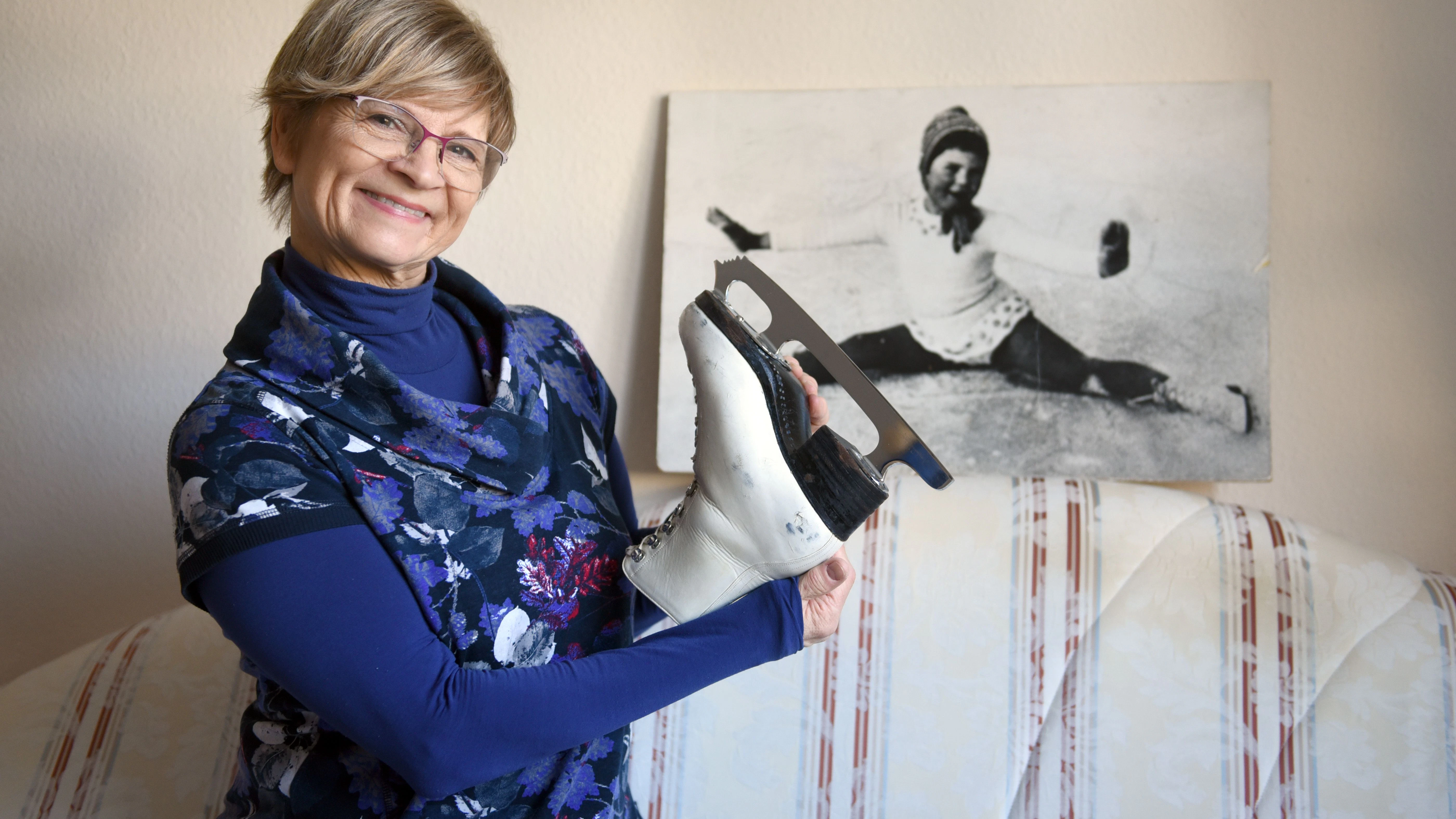 DDR-Eisstar Christine Stüber-Errath steht mit einem alten Schlittschuh vor einem großen Foto, das sie als Fünfjährige zeigt, als sie mit dem Eiskunstlaufen anfing.