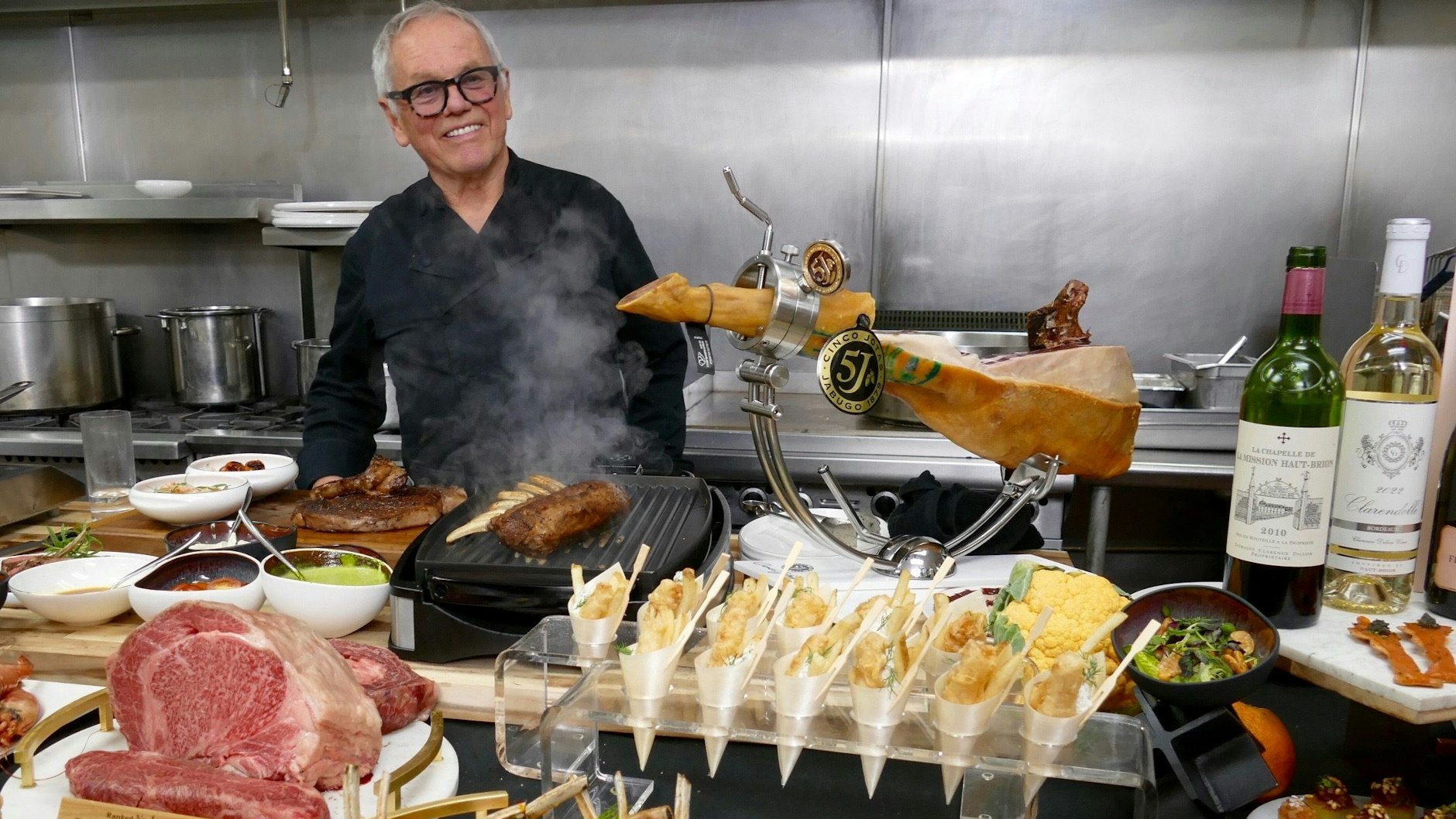 Star-Koch Wolfgang Puck steht neben diversen Gerichten und Zutaten in einer Küche.