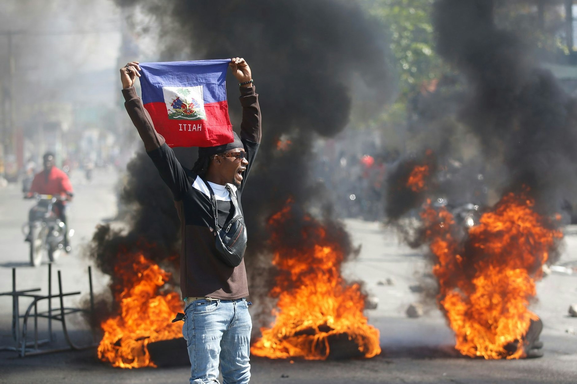 Ein Demonstrant hält eine haitianische Flagge hoch, während hinter ihm Barrikaden brennen.&nbsp;