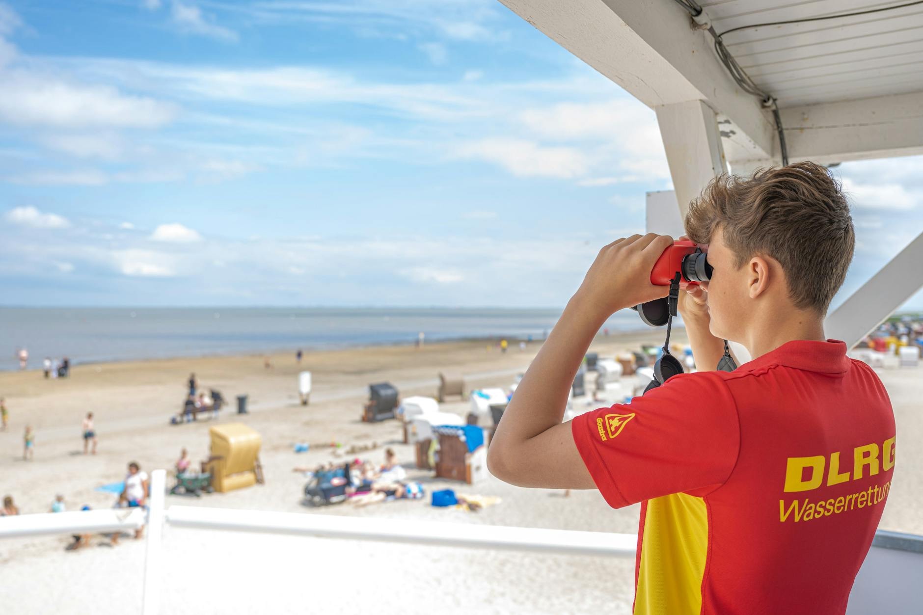 Peter von der DLRG-Wasserrettung steht auf einem Turm und beaufsichtigt mit einem Fernglas den Badestrand im niedersächsischen Dangast. Gesucht werden noch Rettungsschwimmer für die Sommersaison. 