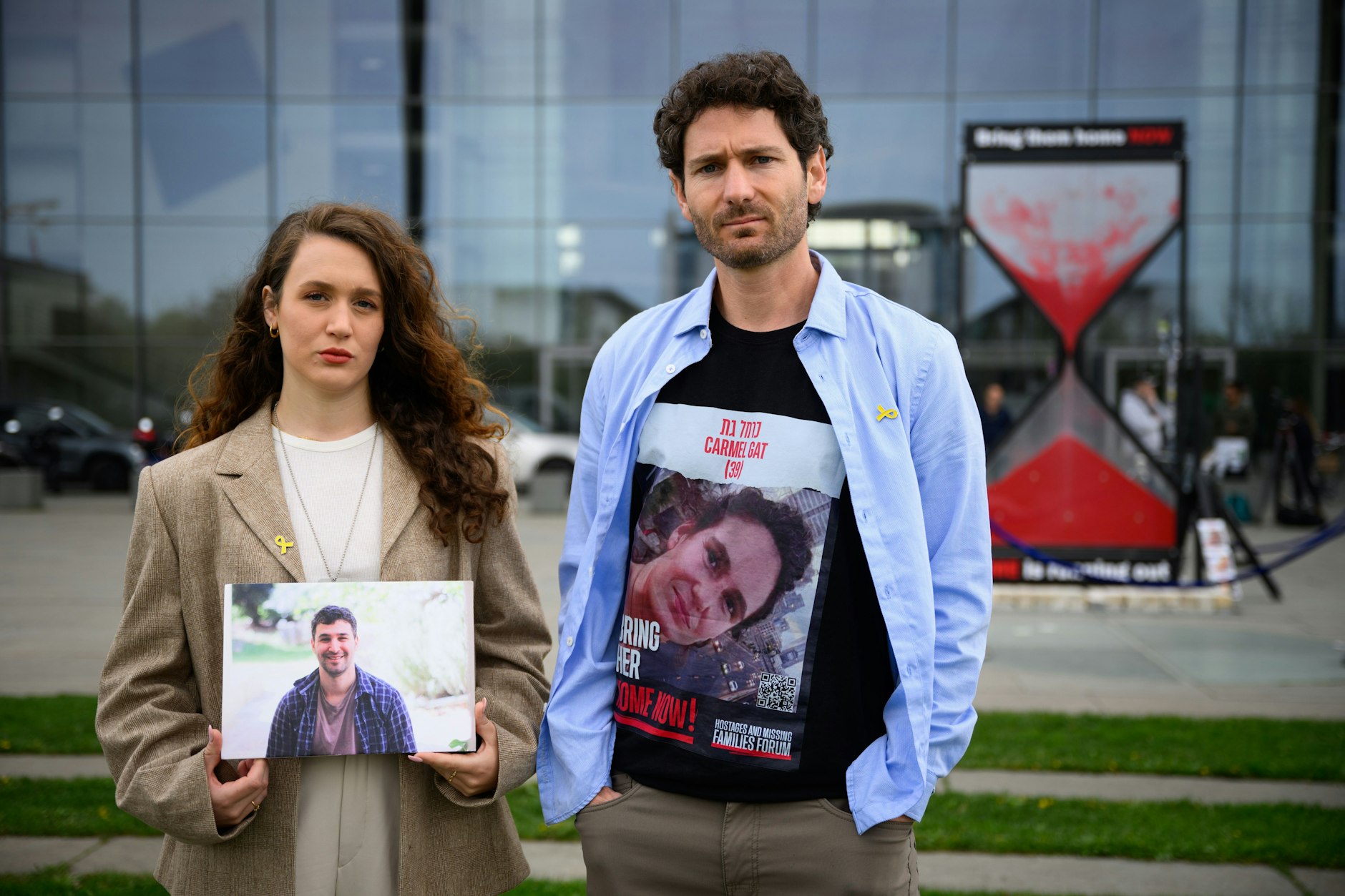 Naama Weinberg (l.), Cousine des in Geiselhaft ermordeten Itai Svirsky, und Alon Gat, Bruder der noch entführten Geisel Carmel Gat und Ehemann der inzwischen befreiten Geisel Yarden Roman-Gat, stehen vor einer mit rotem Sand gefüllten symbolischen Sanduhr und der Aufschrift „Die Zeit läuft davon“ und „Bring them home now“.