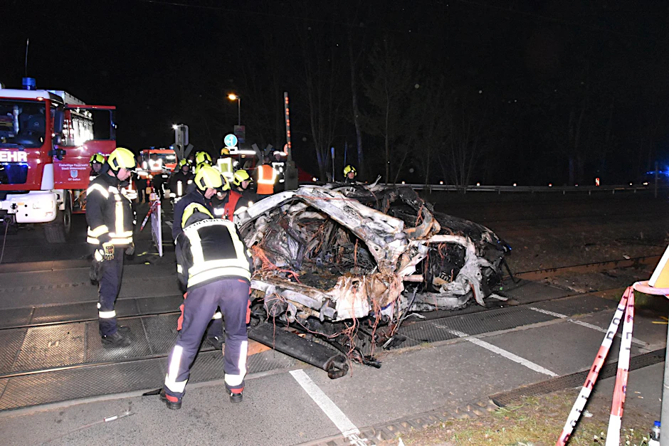 Rettungskräfte stehen am Wrack eines zerstörten Autos nach einem Unfall an einem Bahnübergang.  