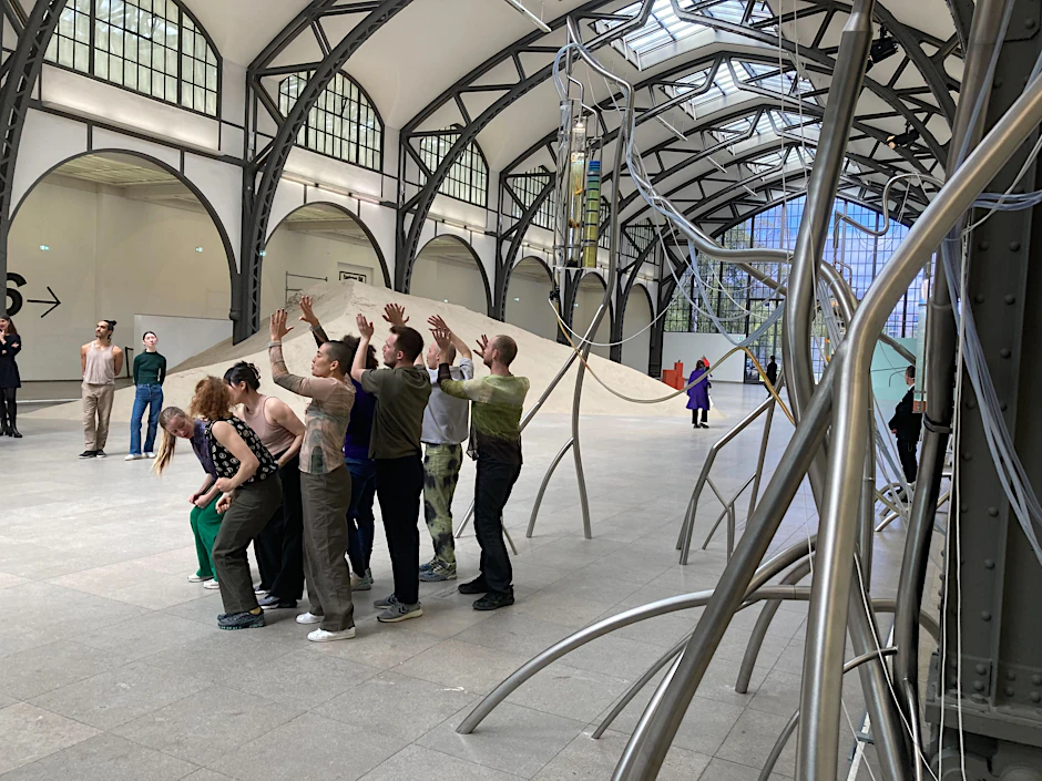 Eine Gruppe Menschen bewegt sich als Teil des Werkes „Attune“ in der Halle der Nationalgalerie der Gegenwart des Hamburger Bahnhofs vor einer aufgeschütteten Sanddüne.&nbsp;