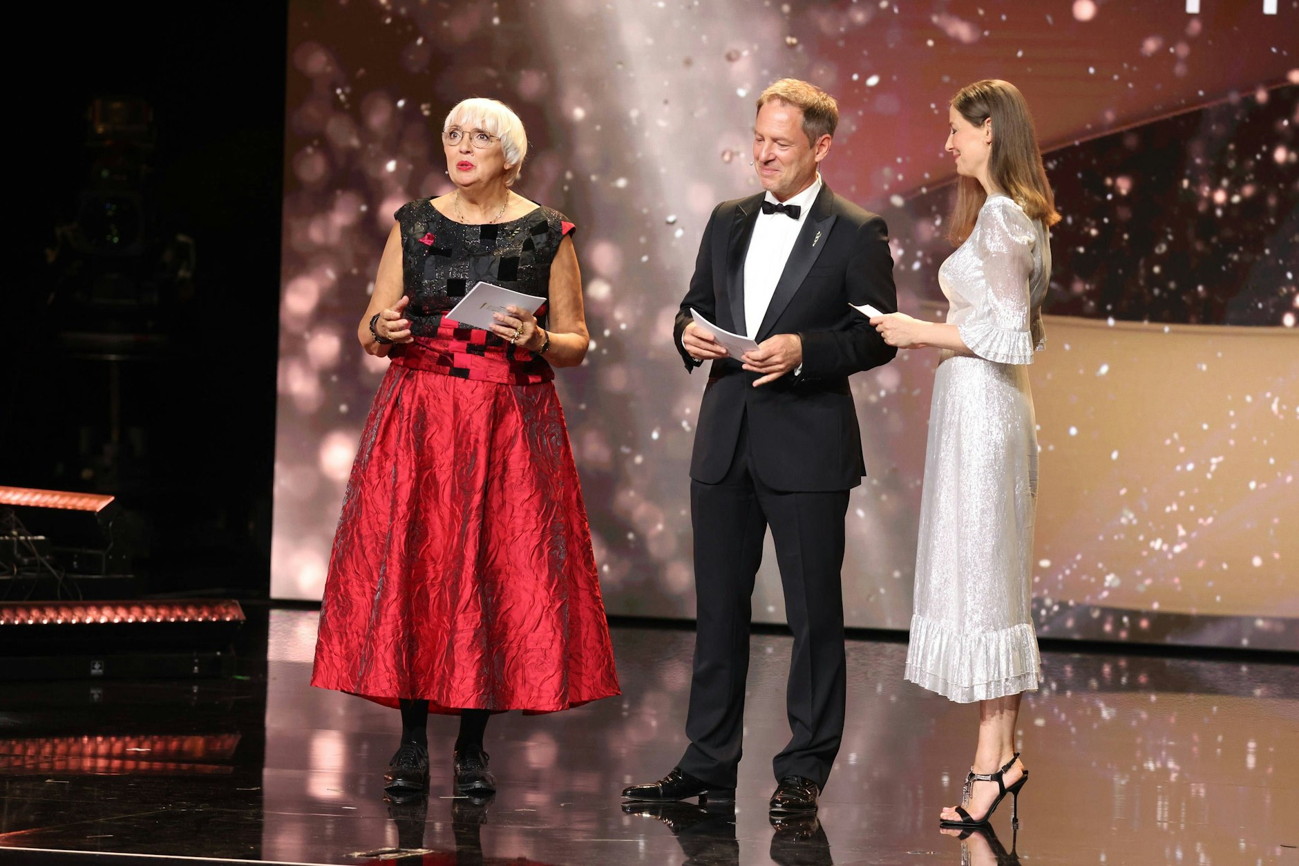 <strong>TOP/FLOP:</strong>&nbsp;<strong>Claudia Roth</strong> (l.), <strong>Florian Gallenberger</strong> und <strong>Alexandra Maria Lara</strong> auf der Bühne. Wir sind begeistert von Roths roter Robe und entschuldigen uns für diese müde Alliteration. Auch Florian Gallenberger hat alles richtig gemacht. Alexandra Maria Laras Kleid indes kann sich nicht entscheiden zwischen Gala und Picknick. Die silbrige Faser sieht nach Abendkleid aus, Länge und die trutschigen Rüschen hingegen erinnern mehr an einen Sommerausflug mit dem Hollandrad.