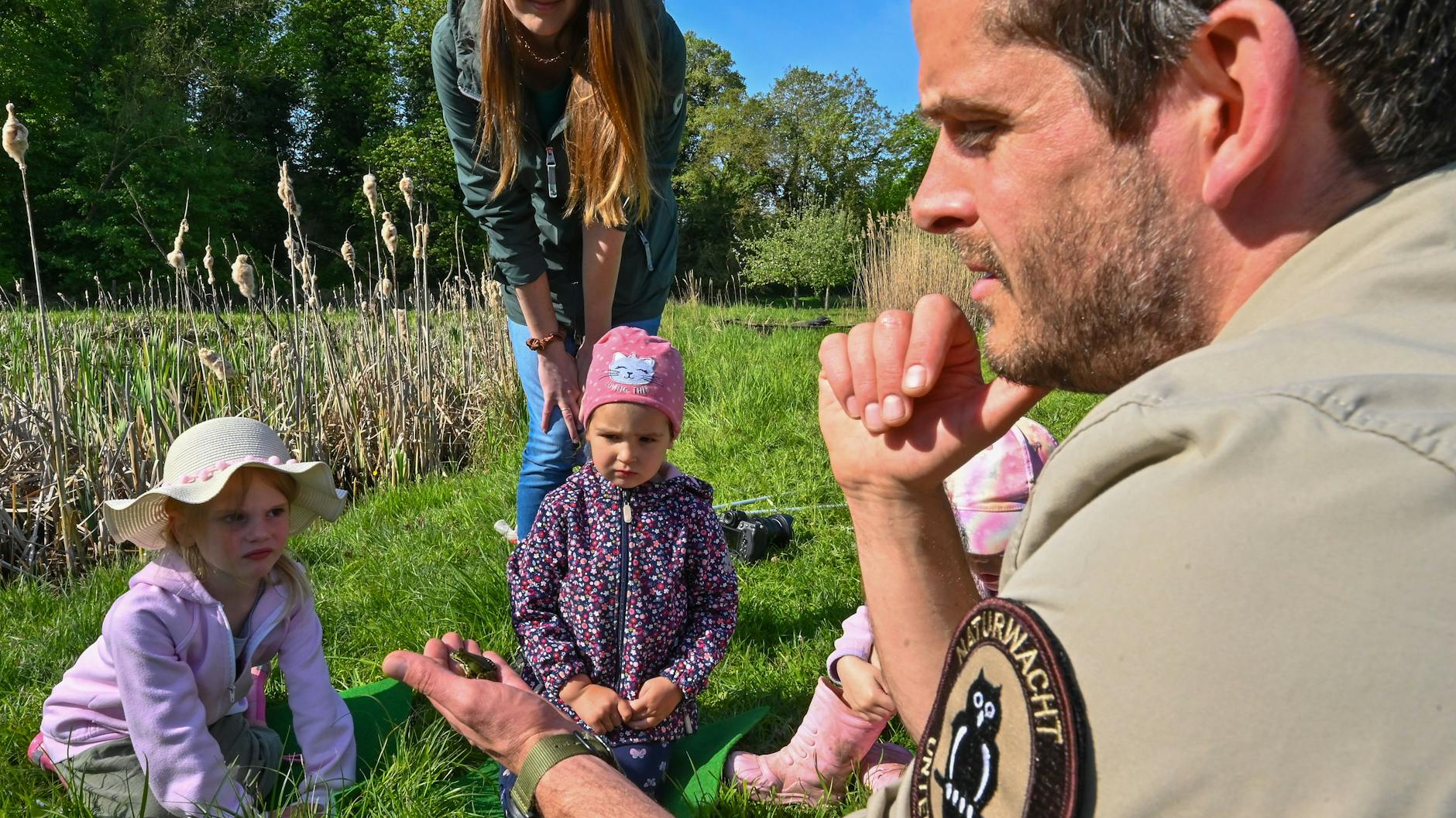 Andreas Hein, Ranger der Naturwacht Brandenburg, erklärt Kindern der Kita „Kleine Wildhüter“ das Leben von Amphibien auf einer Wiese im Nationalpark Unteres Odertal. Julia Werner, Leiterin der Kita, schaut zu.