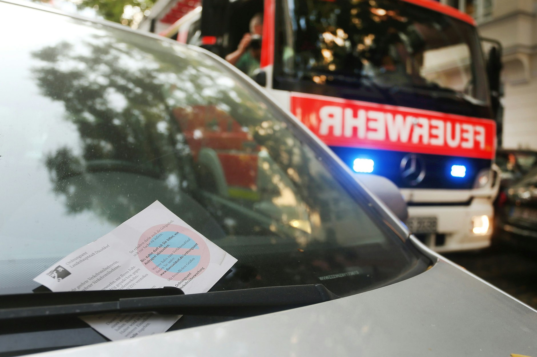 Ein falsch geparktes Auto mit Knöllchen, im Hintergrund ein Feuerwehrauto mit Blaulicht.