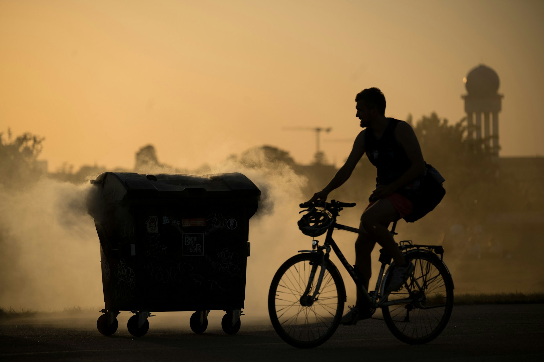 Ein Fahrradfahrer fährt vor Sonnenuntergang auf dem Tempelhofer Feld an einem qualmenden Mülleimer nahe dem Grillplatz vorbei.