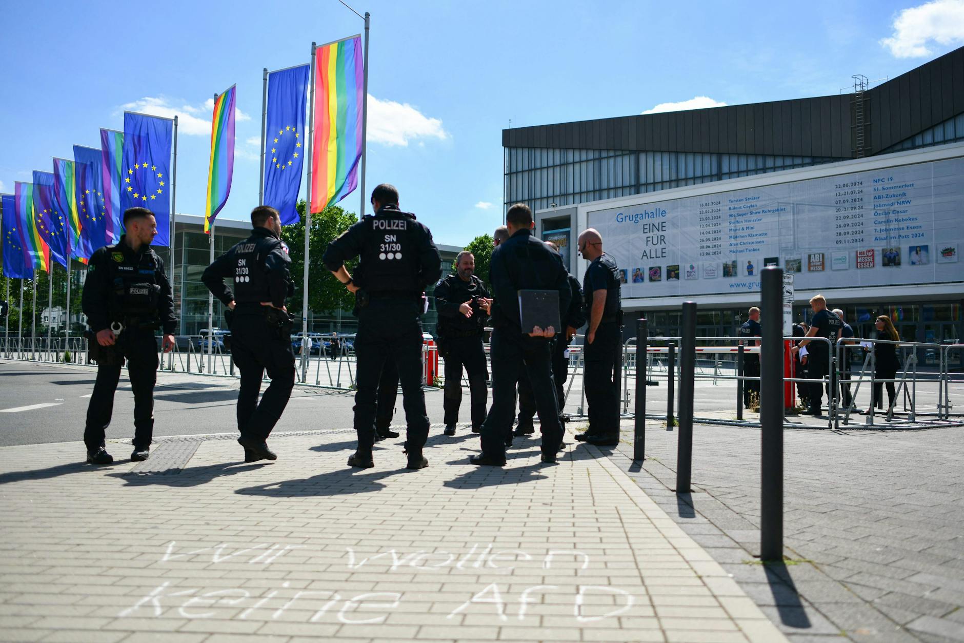 Polizisten stehen vor der Grugahalle in Essen, wo der AfD-Bundesparteitag stattfinden soll. EU- und Regenbogen-Fahnen wehen im Wind. Auf dem Gehweg steht: „Wir wollen keine AfD“. 