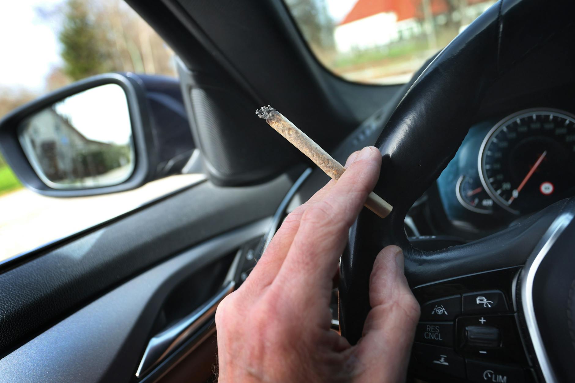 Ein Mann sitzt mit einem Joint zwischen den Fingern am Steuer eines Autos. Brandenburgs Innenminister Michael Stübgen hat den Wirkstoff-Grenzwert für Cannabis am Steuer kritisiert. (Symbolbild) 