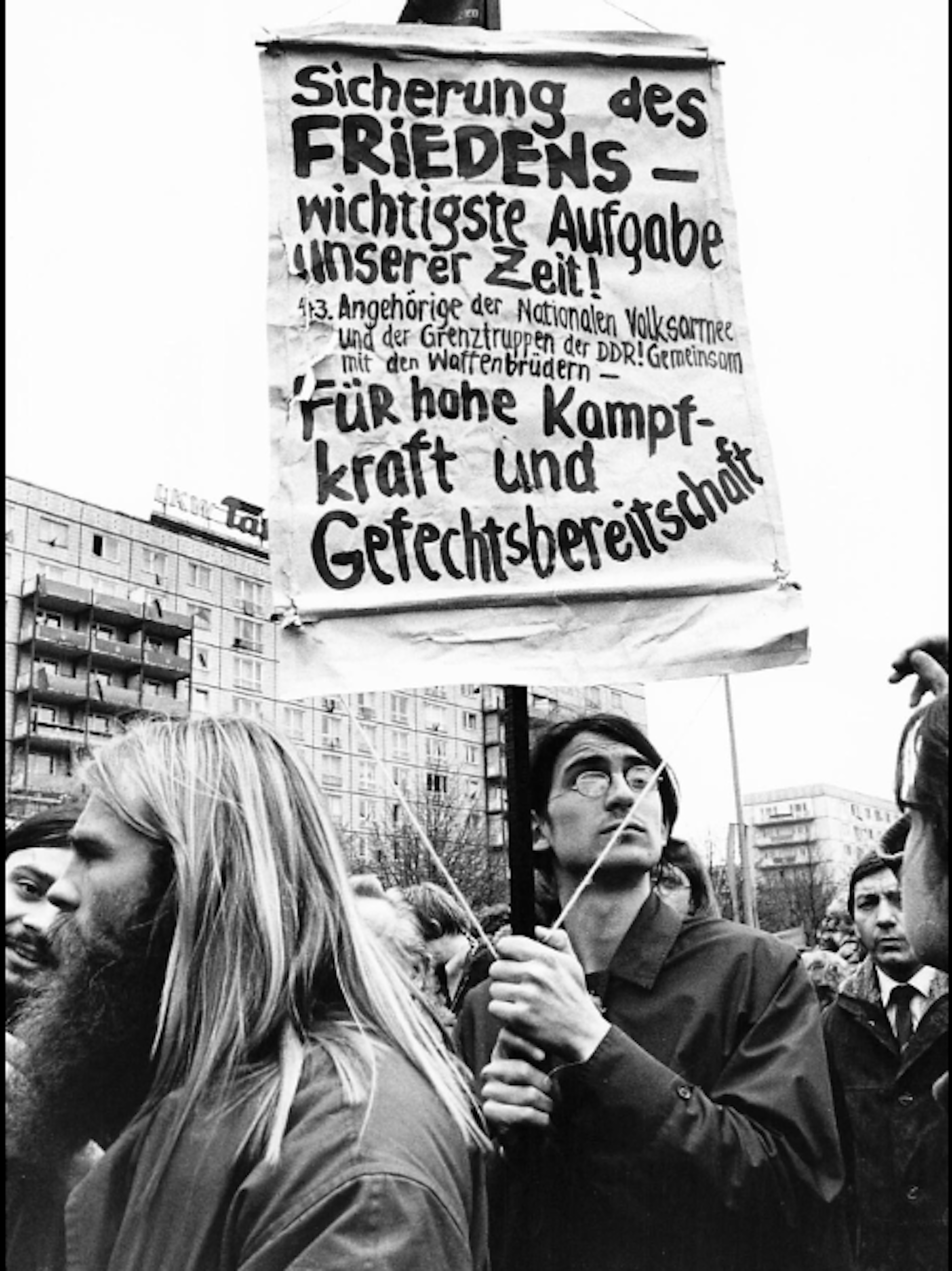 1.-Mai-Demo in Ost-Berlin: Hier wird ein nicht ganz ernst gemeintes Plakat gezeigt, auf dem gleich mehrere Kampfparolen stehen, die Tage zuvor in DDR-Zeitungen vorgegeben wurden.