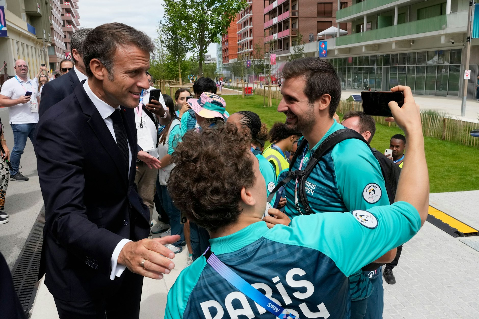 Der französische Präsident Emmanuel Macron posiert für ein Selfie mit freiwilligen Olympia-Helfern, als er das Olympische Dorf besucht. Erste Helfer verkaufen jetzt schon ihre Klamotten auf ebay und Co.