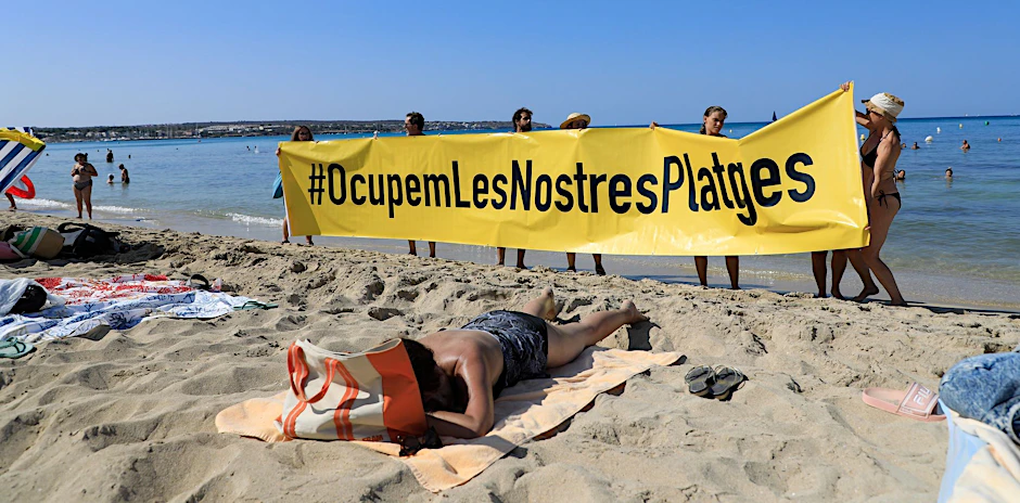 Leute halten an einem Strand auf Mallorca ein Banner mit der Schrift „#Besetzen wir unsere Strände“ aus Protest gegen den Massentourismus auf der Balearen-Insel.&nbsp;