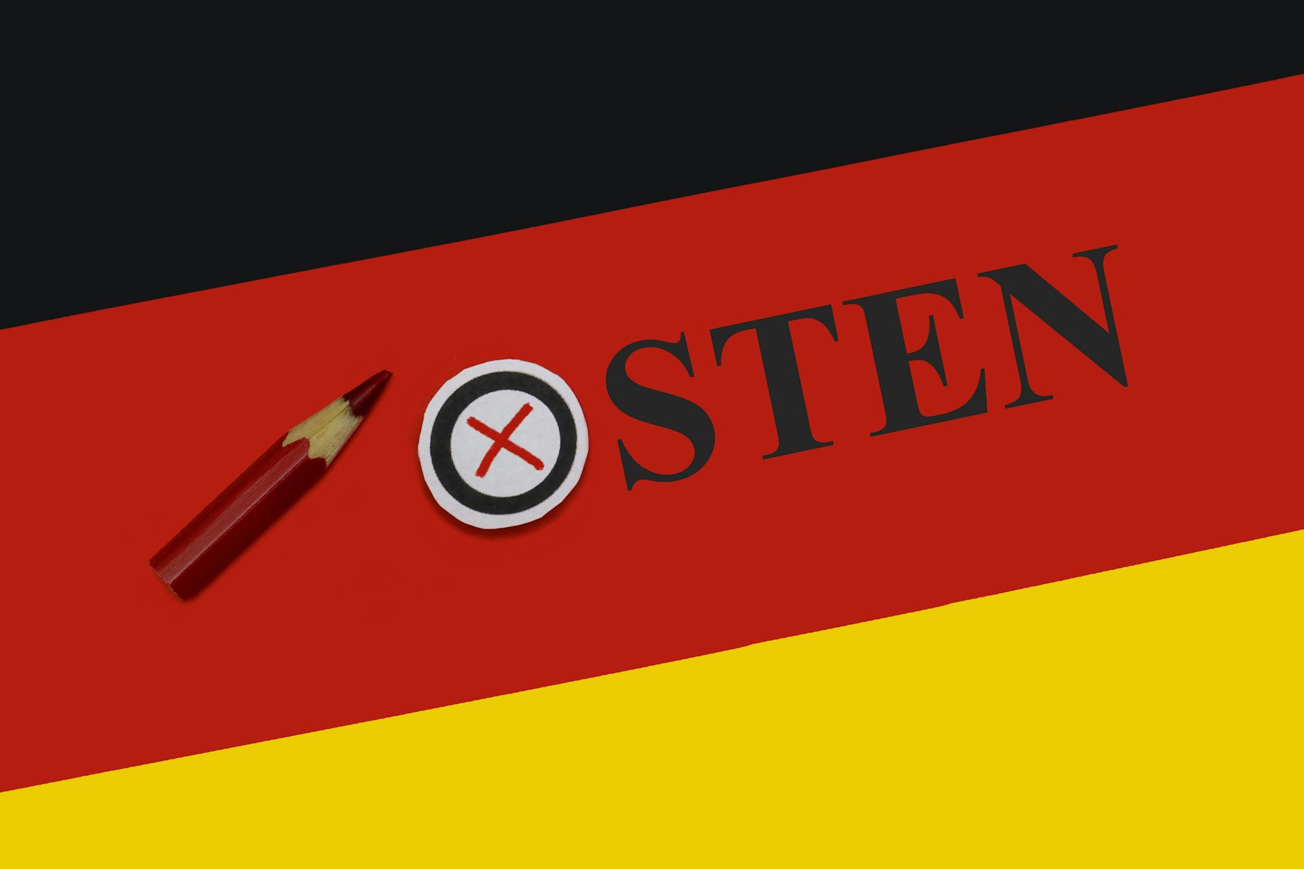 Auf dem roten Streifen der Deutschlandfahne steht Osten und im O ist mit Rot ein Wahlkreuz gesetzt. 