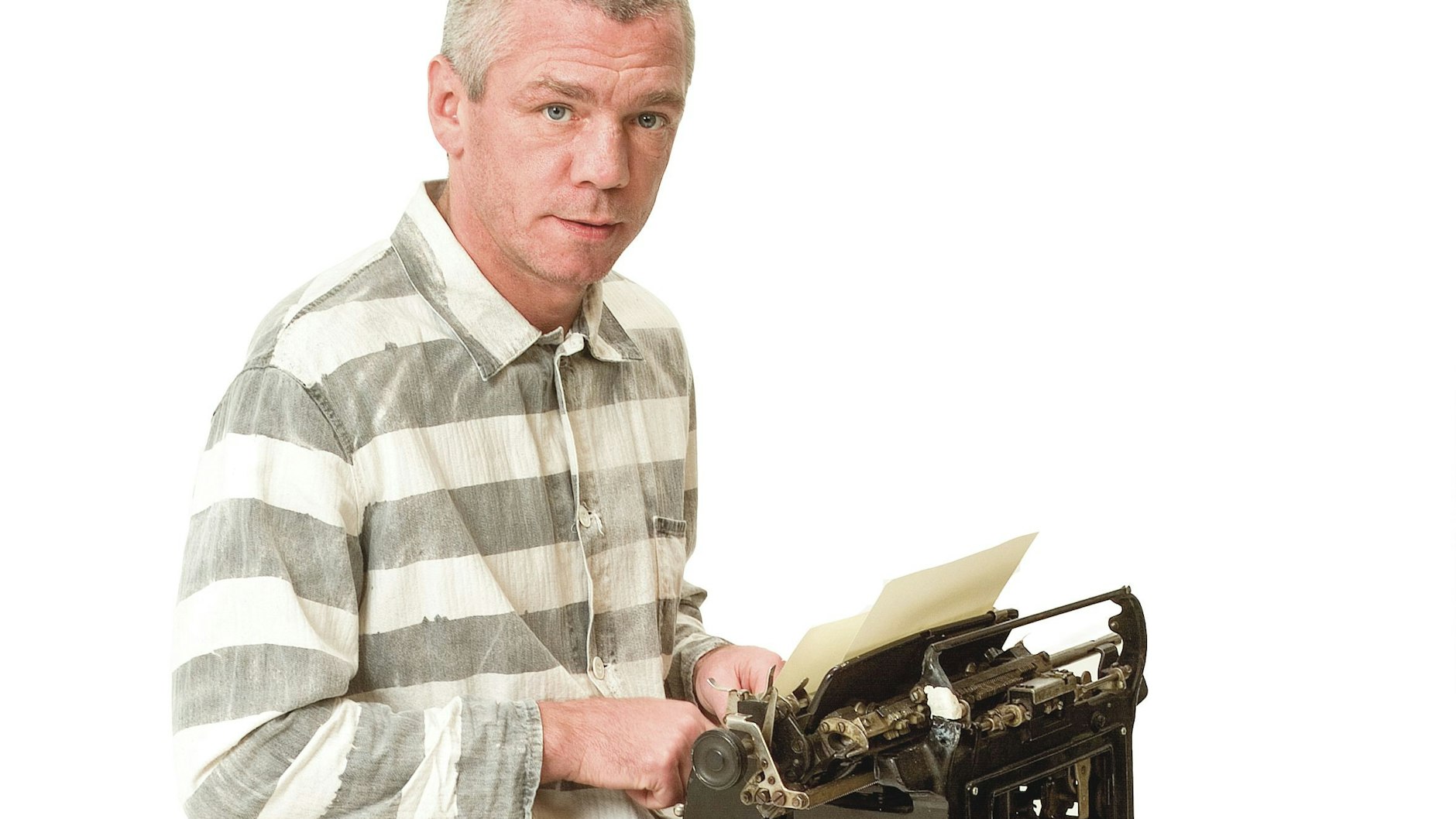 Graciano Rocchigiani sitzt in der Verkleidung eines Häftlings auf einem Schemel, hat eine Schreibmaschine auf dem Schoß.