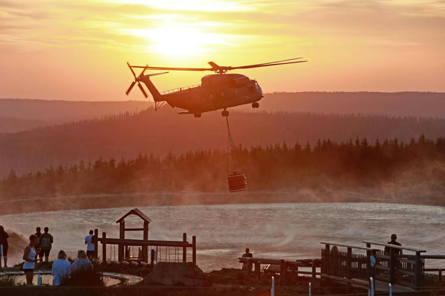 Ein Hubschrauber der Bundeswehr fliegt im Licht der untergehenden Sonne mit einem Wasserbehälter zum Königsberg am Brocken.