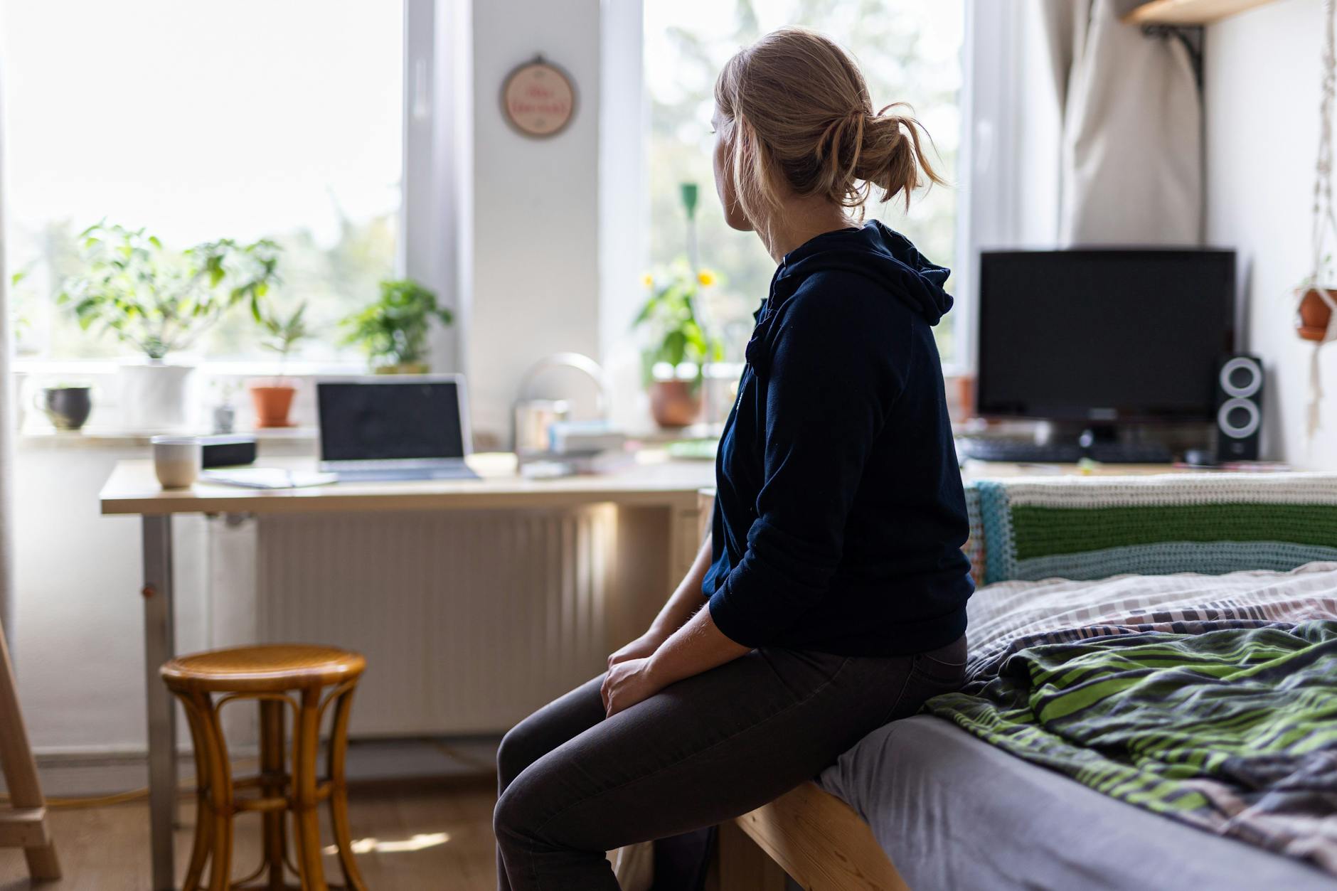Eine Studentin sitzt auf dem Bett in ihrem WG-Zimmer und schaut aus dem Fenster.