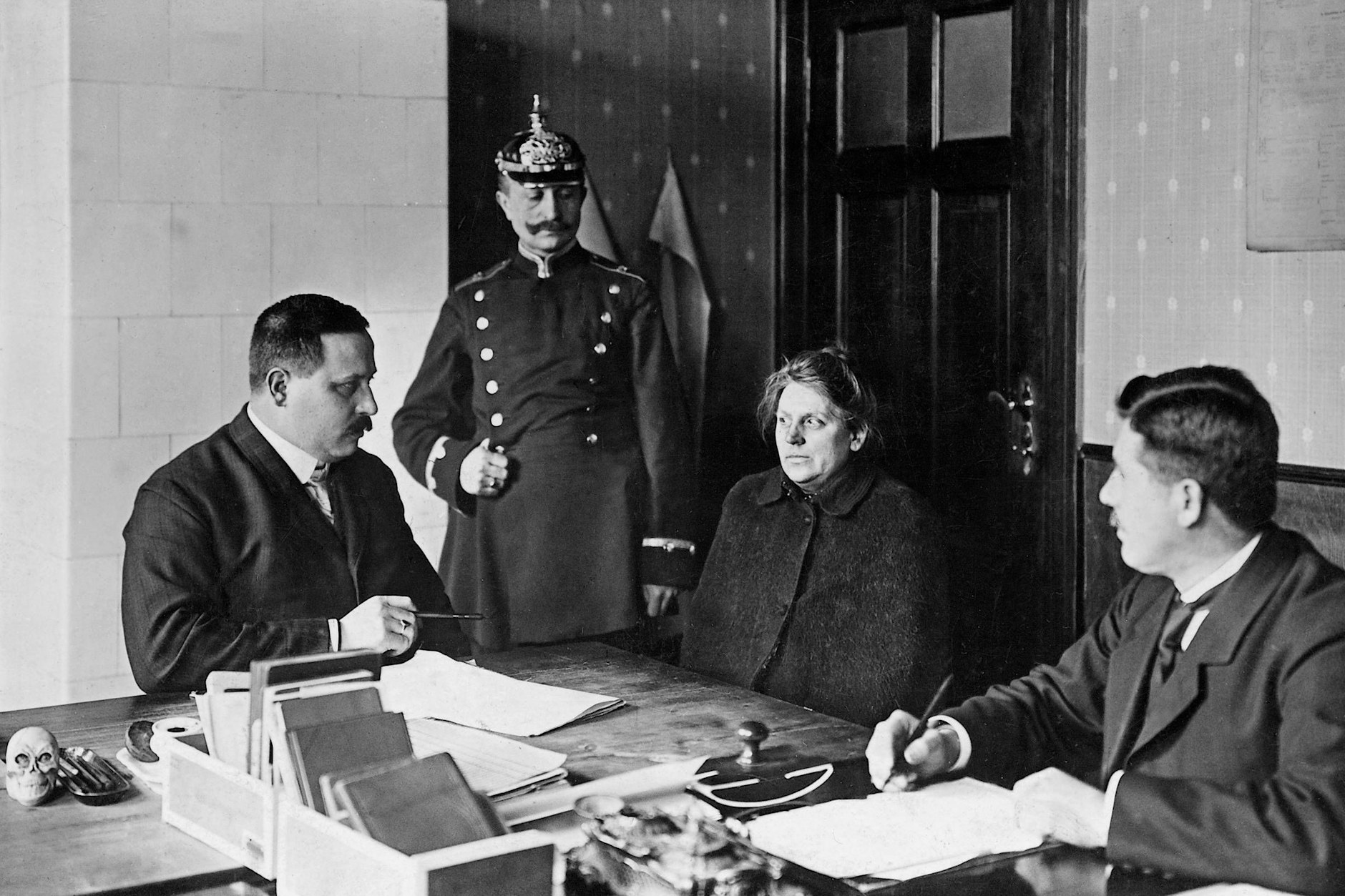 Auf einem Polizeirevier wird eine Frau von zwei Polizeibeamten in Zivil befragt. Ein Polizist in Uniform steht daneben. Das Foto stammt aus dem Jahr 1911.