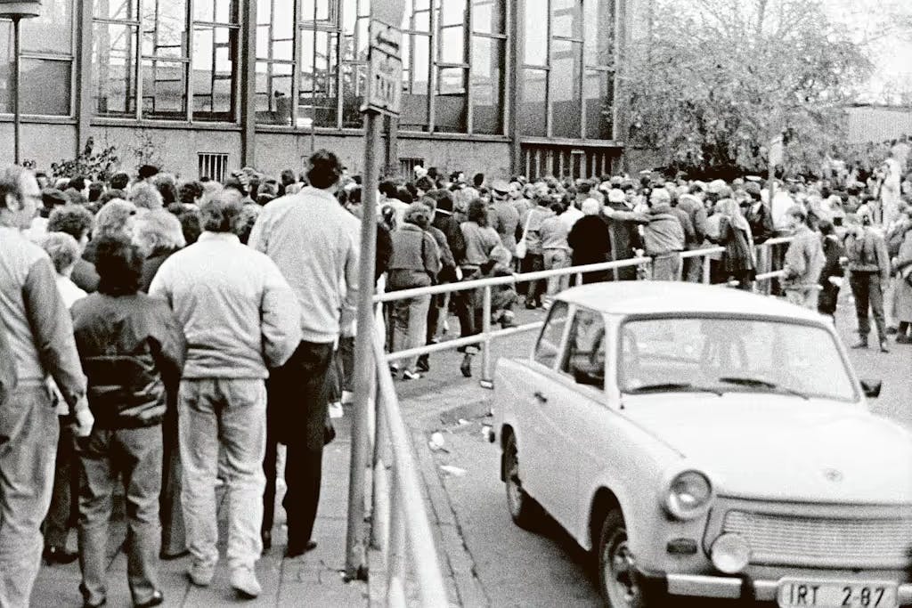 Am 10. November 1989 steht eine Menschenmenge dichtgedrängt vor dem Tränenpalast am Bahnhof Friedrichstraße und will in den Westteil Berlins. Im Halteverbot steht ein Trabi, der nun Fragen aufwirft.