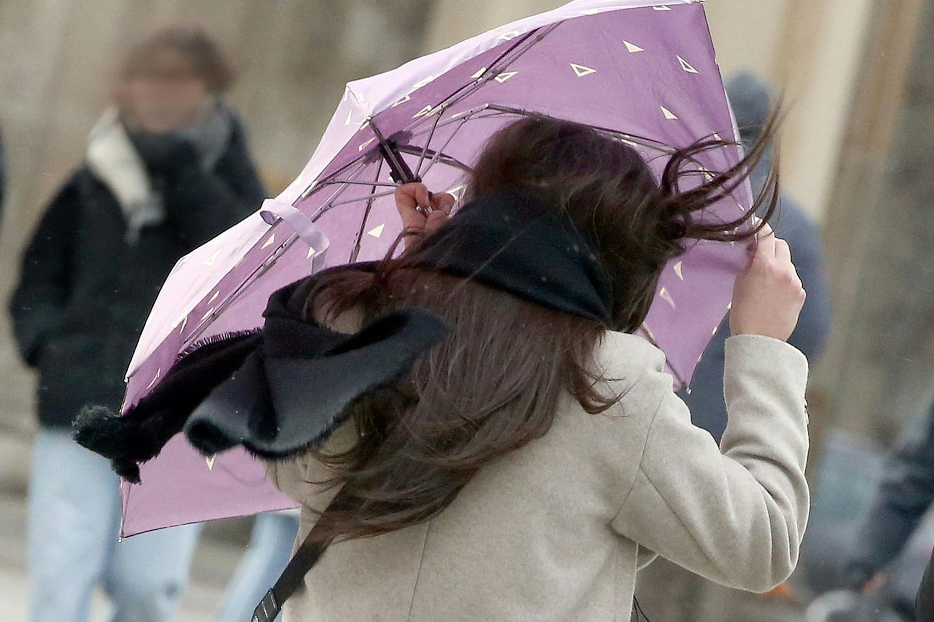 Eine Passantin mit wehenden Haaren sichert ihren Regenschirm, während sie durch Berlin läuft.