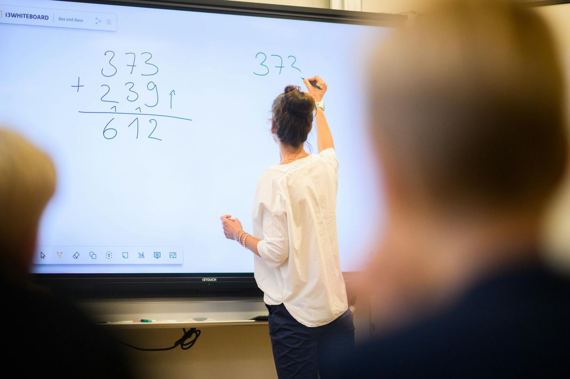 Eine Lehrerin schreibt eine Mathematikaufgabe auf eine digitale Schultafel im Klassenraum einer 4. Klasse einer Grundschule.