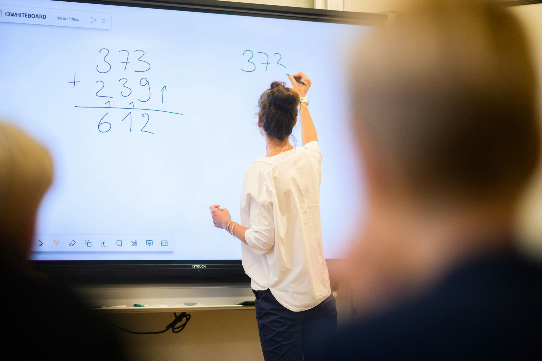 Eine Lehrerin schreibt eine Mathematikaufgabe auf eine digitale Schultafel im Klassenraum einer 4. Klasse einer Grundschule.