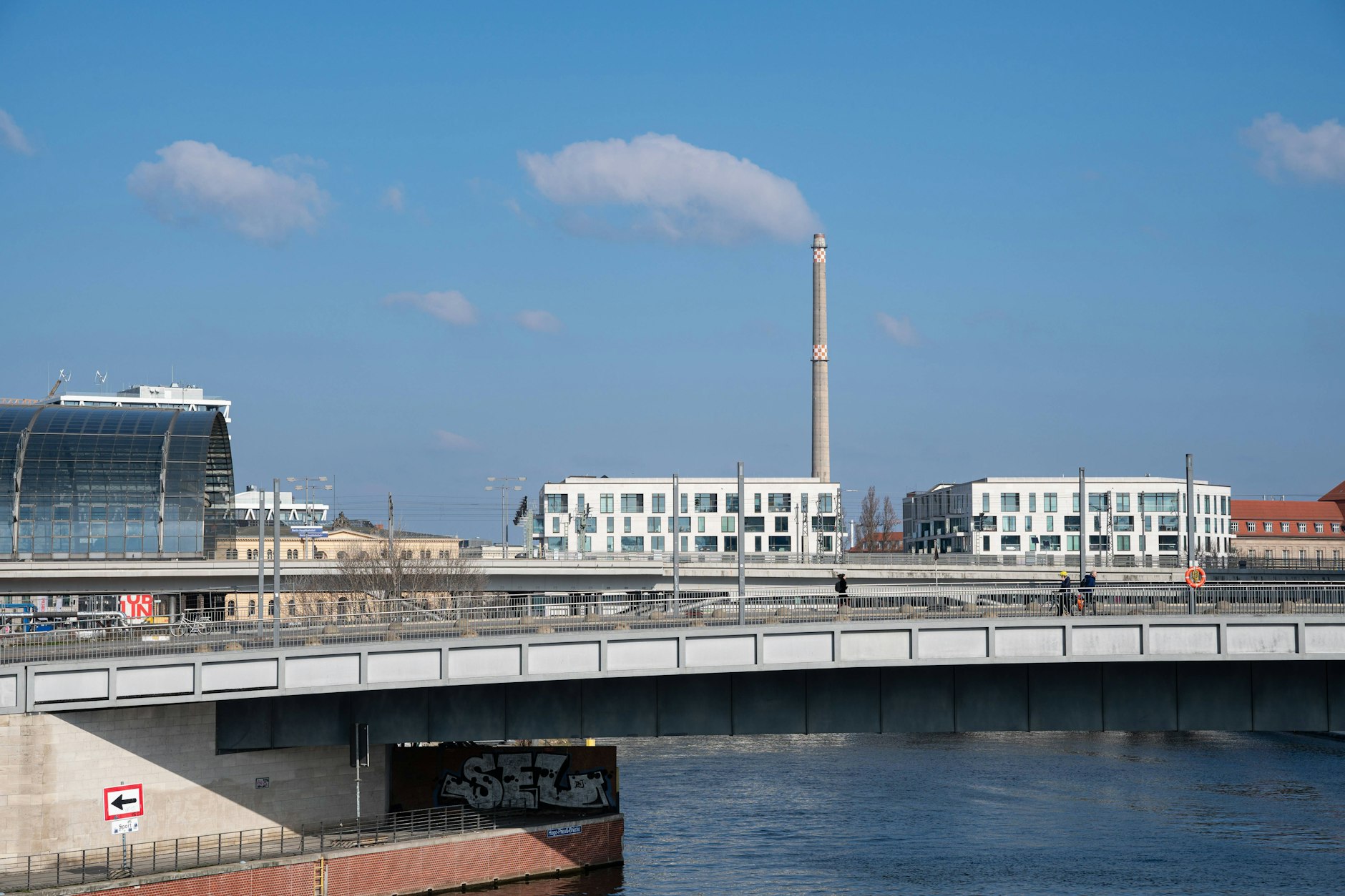 Vom Spreebogenpark aus öffnet sich der Blick auf den Humboldthafen mit der Hugo-Preuß-Brücke und der Humboldthafenbrücke. Dahinter ragt der östliche Teil des Berliner Hauptbahnhofs empor, während im Hintergrund der 150 Meter hohe Schornstein des Heizkraftwerks Scharnhorststraße noch die Skyline ziert – eingefangen an einem sonnigen Frühlingstag.