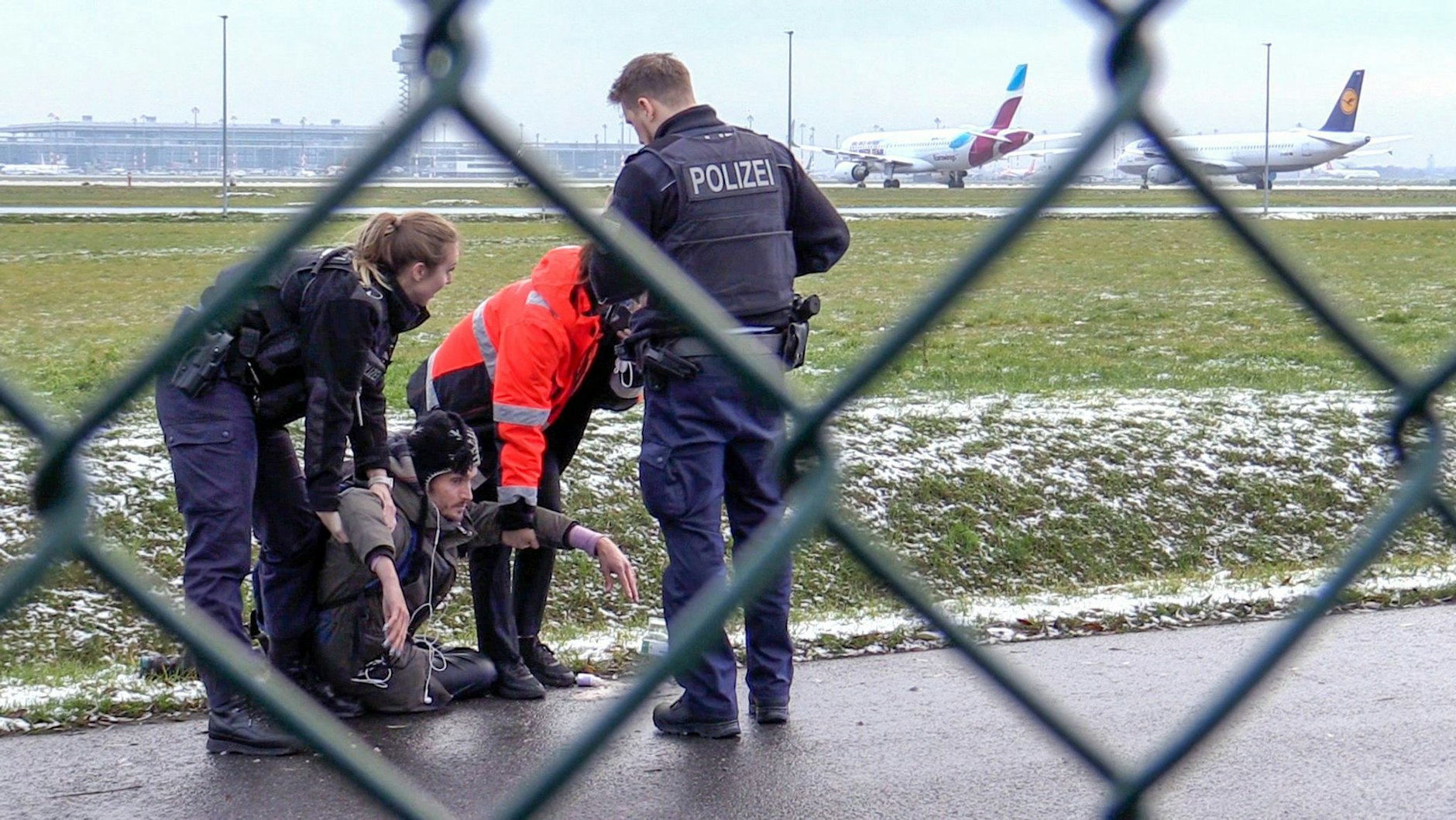 Zwei Beamte der Bundespolizei und ein Sicherheitsmitarbeiter nehmen einen Klimakleber nahe der Start- und Landebahn des BER fest (2022).