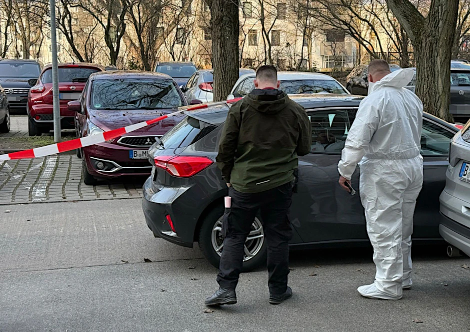 Zwei Beamte der Spurensicherung stehen vor einem Auto, das mit Absperrband gesichert ist.