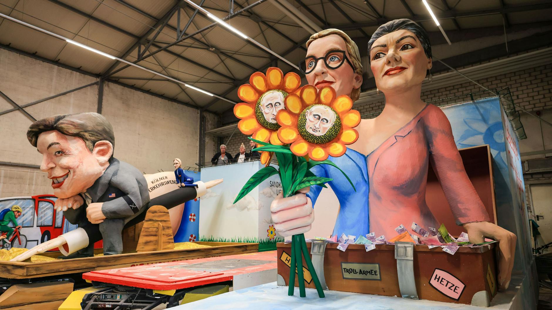 Zweimal Alice Weidel: Links winzig zusammen mit einem mächtigen Elon Musk auf einem Hitlergruß-Arm als Wippe. Rechts: Alice Weidel und Sahra Wagenknecht als Putin-freundliches Duo. Ein Motivwagen des Kölner Rosenmontagszuges in der Wagenhalle.