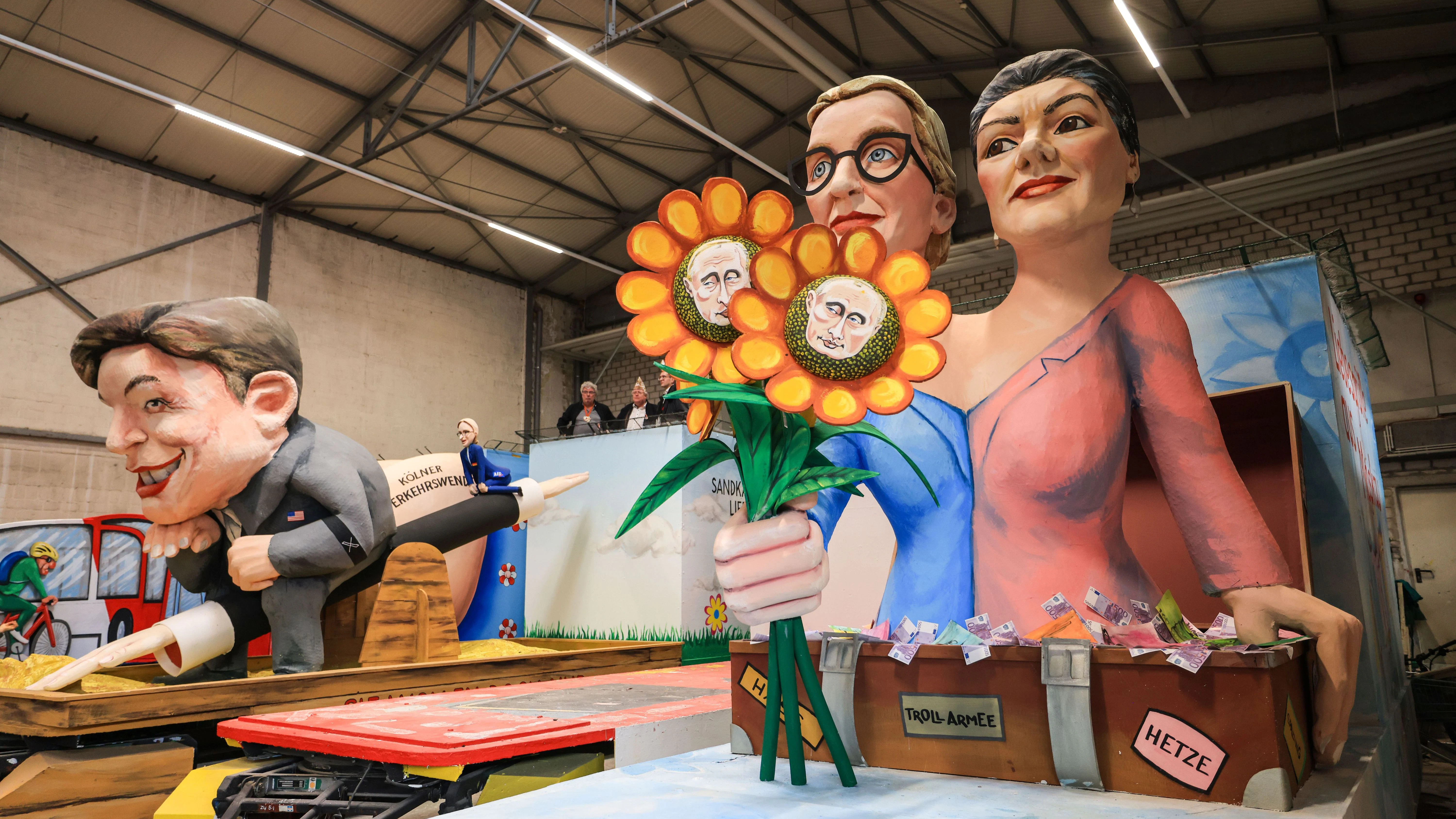 Zweimal Alice Weidel: Links winzig zusammen mit einem mächtigen Elon Musk auf einem Hitlergruß-Arm als Wippe. Rechts: Alice Weidel und Sahra Wagenknecht als Putin-freundliches Duo. Ein Motivwagen des Kölner  Rosenmontagszuges in der Wagenhalle.