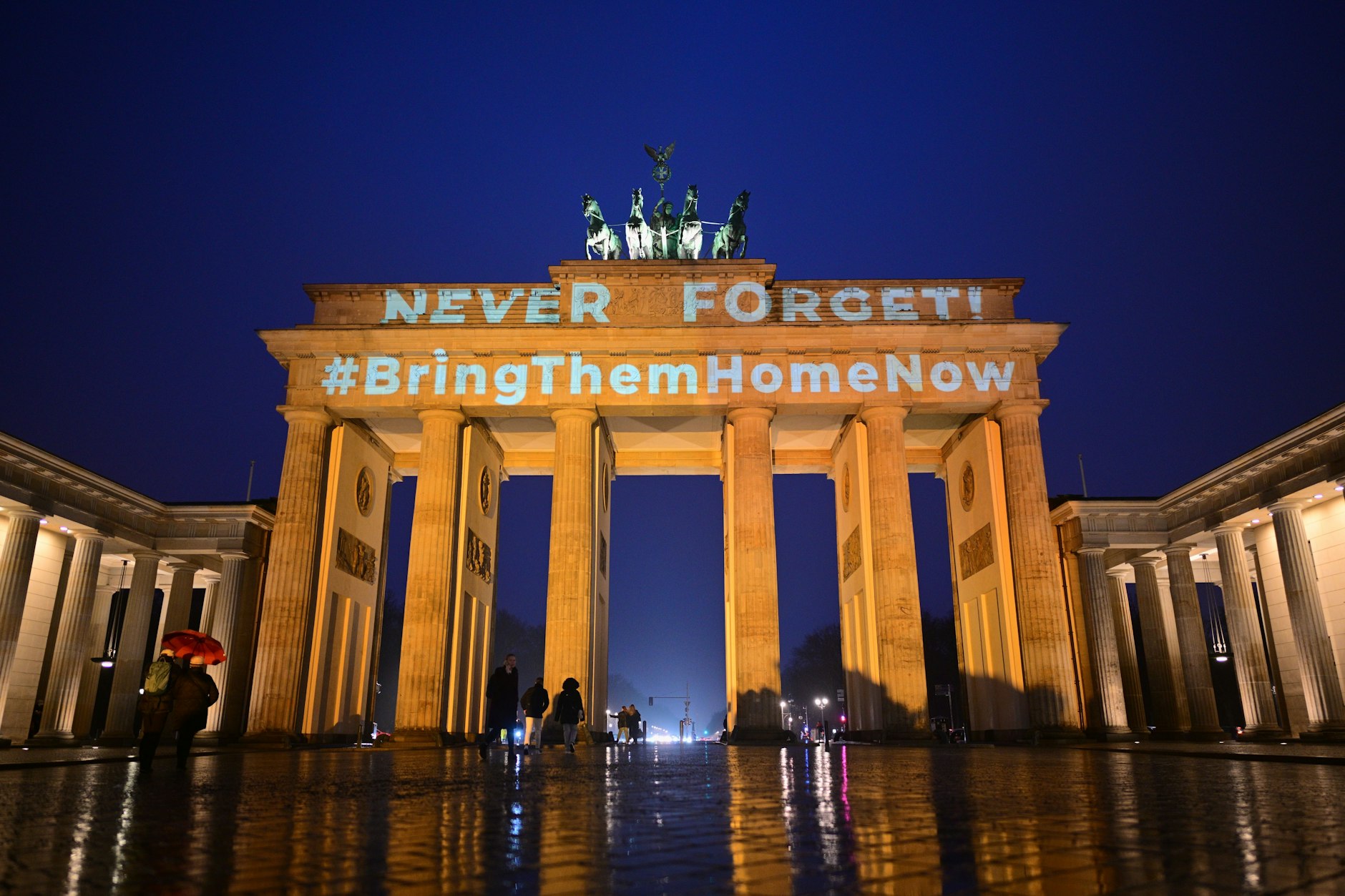 Die Schriftzüge „Never forget“&nbsp;und „#BringThemHomeNow“&nbsp;werden auf das orange angestrahlte Brandenburger Tor projiziert.