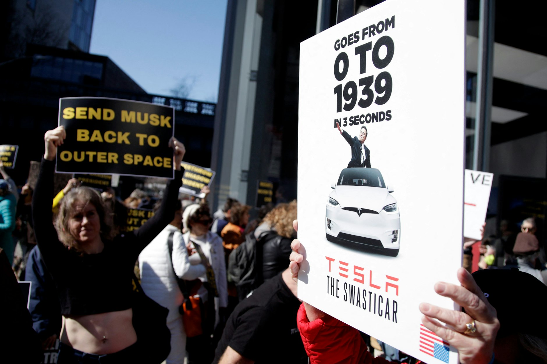 Bei einer Kundgebung gegen Tesla in New York hält ein Demonstrant ein Schild hoch, auf dem Teslas als „Swasticars“ bezeichnet werden. Hinter einem Tesla-Fahrzeug reckt Unternehmenschef Elon Musk die Hand zum Hitlergruß hoch, darüber der zynische Slogan: „von null auf 1939 in drei Sekunden“.