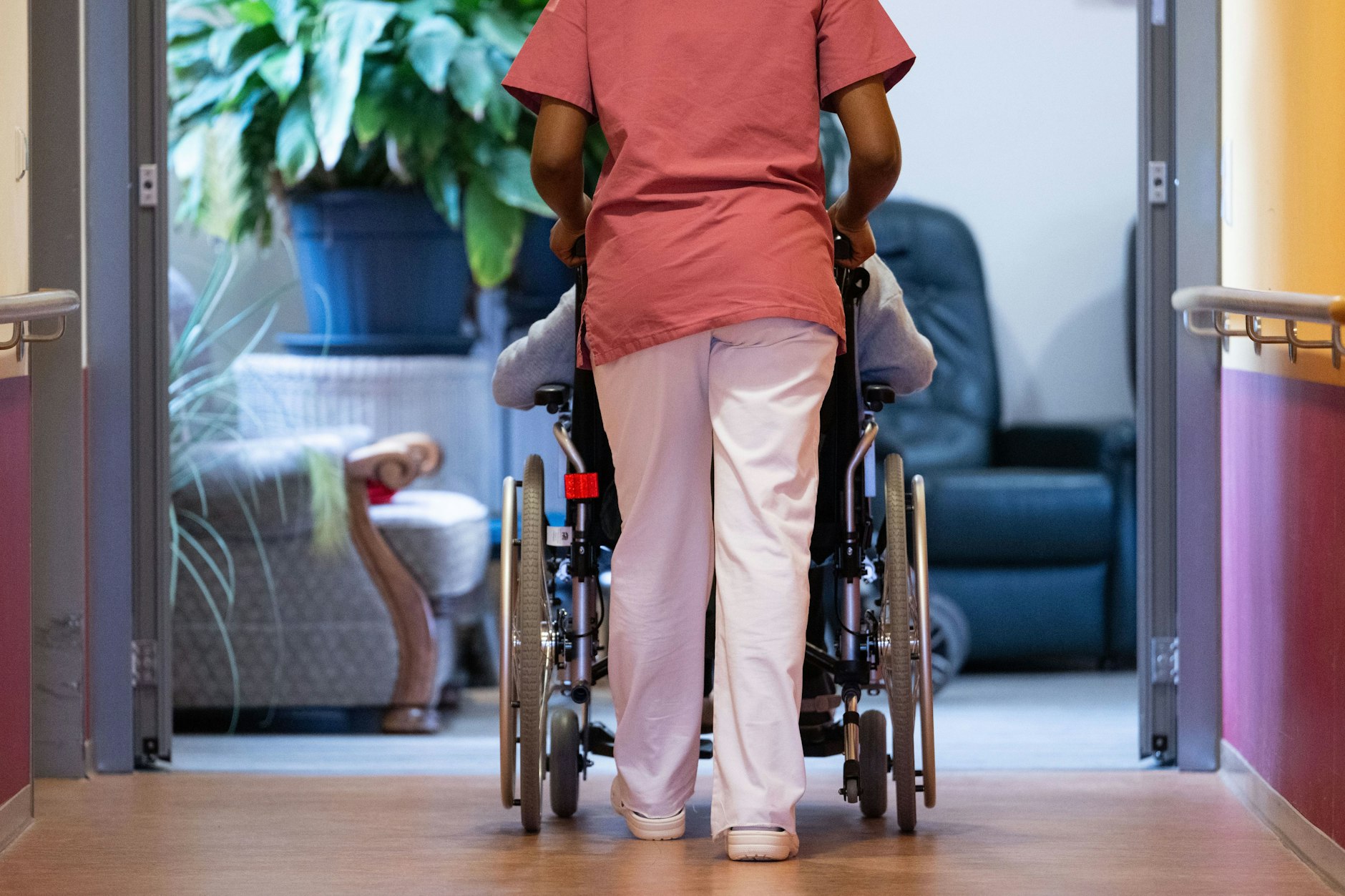 Eine Bewohnerin eines Pflegeheims wird in ihrem Rollstuhl einen Gang entlang geschoben.
