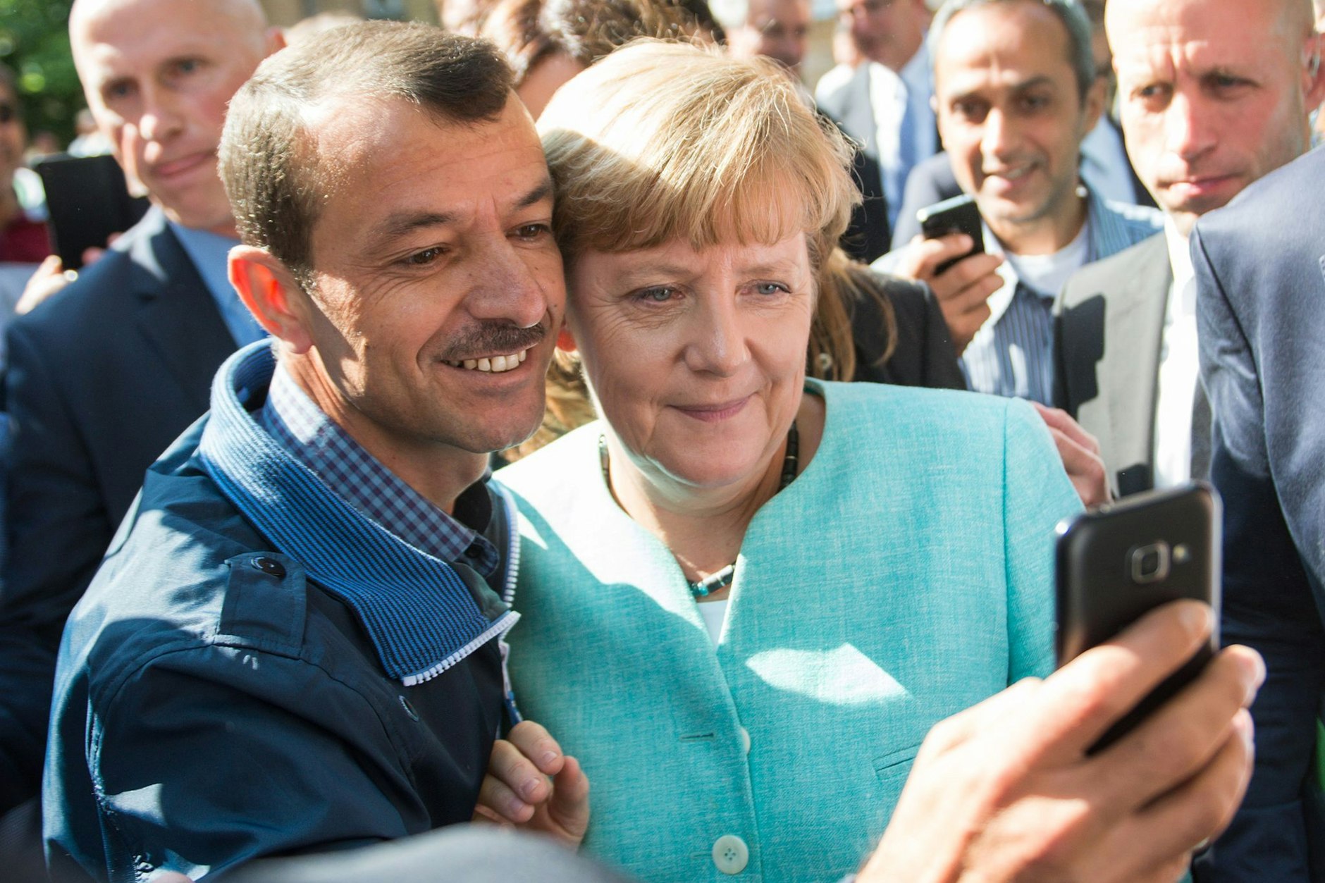 Angela Merkel und ein Mann, der 2015 als Flüchtling nach Deutschland gekommen ist, machen ein Selfie zusammen.