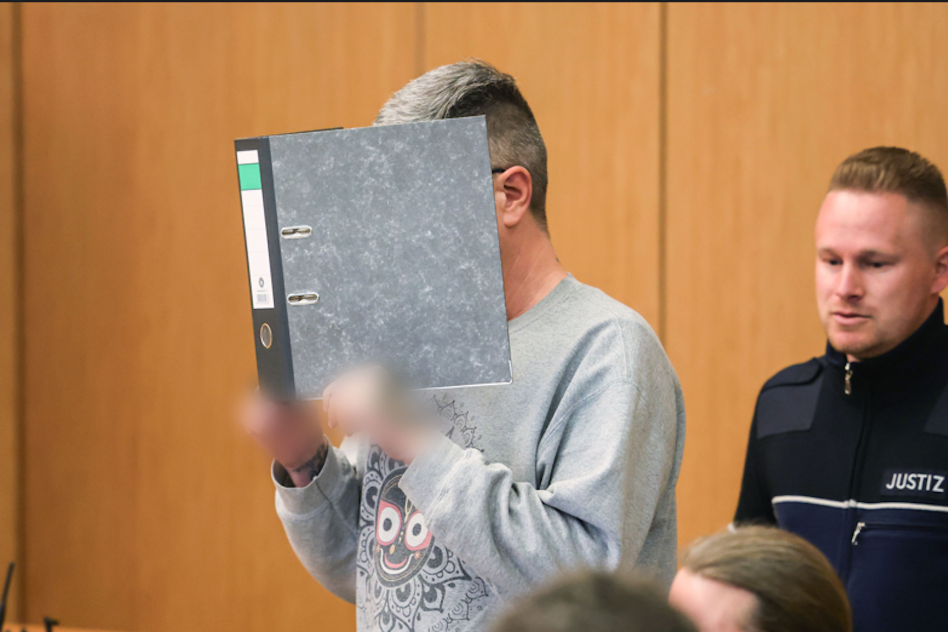 Der wegen 9-fachen Mordes angeklagte Krankenpfleger hält sich im Gerichtssaal in Aachen einen Aktenordner vor das Gesicht. Seine Hände wurden aus rechtlichen Gründen gepixelt.