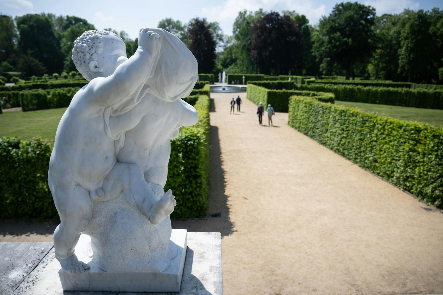Menschen gehen durch die Parkanlage Sanssouci, im Vordergrund steht eine Steinfigur. Die Pflege der Parkanlagen ist aufwendig und teuer.