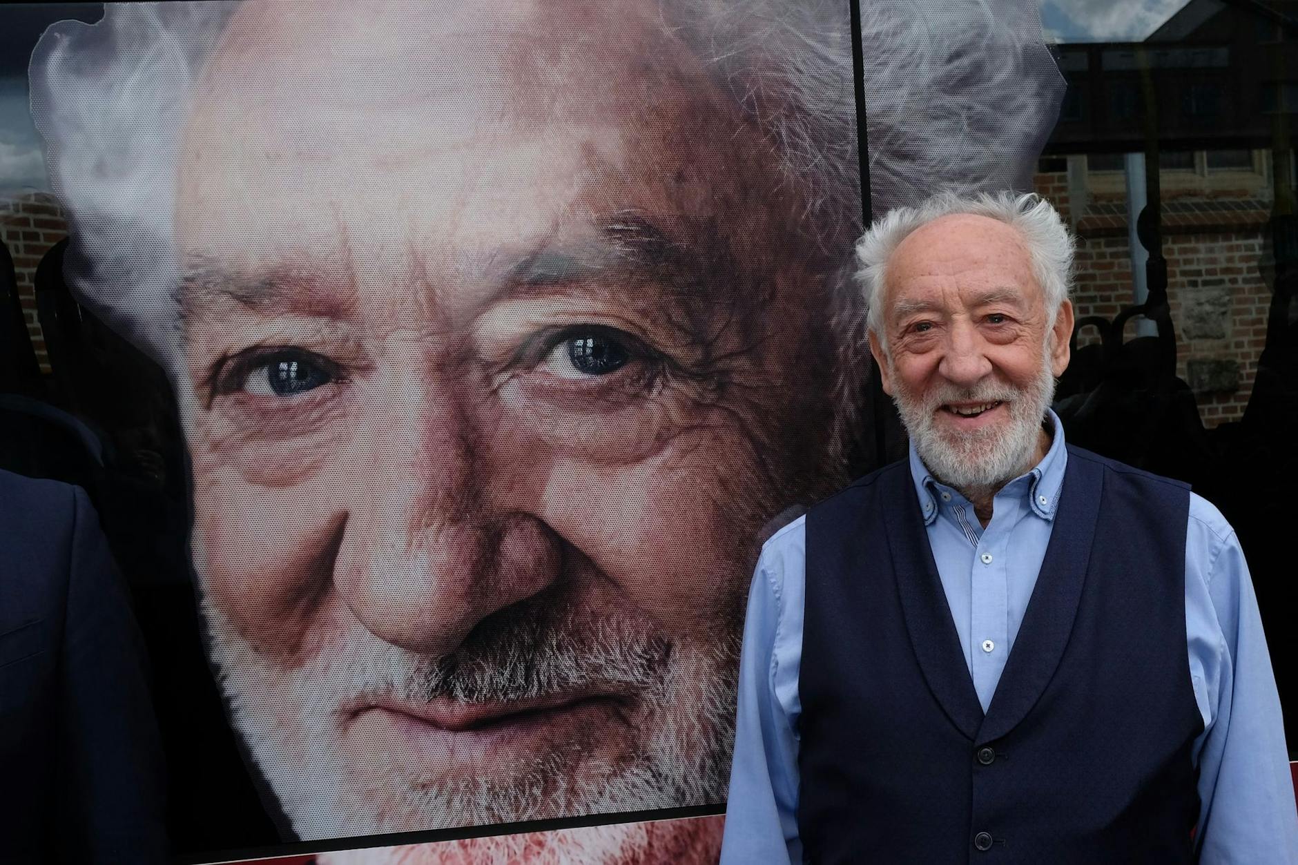 Dieter Hallervorden steht vor einem Porträt seiner selbst, das zu Werbezwecken auf einem Bus zu sehen ist.