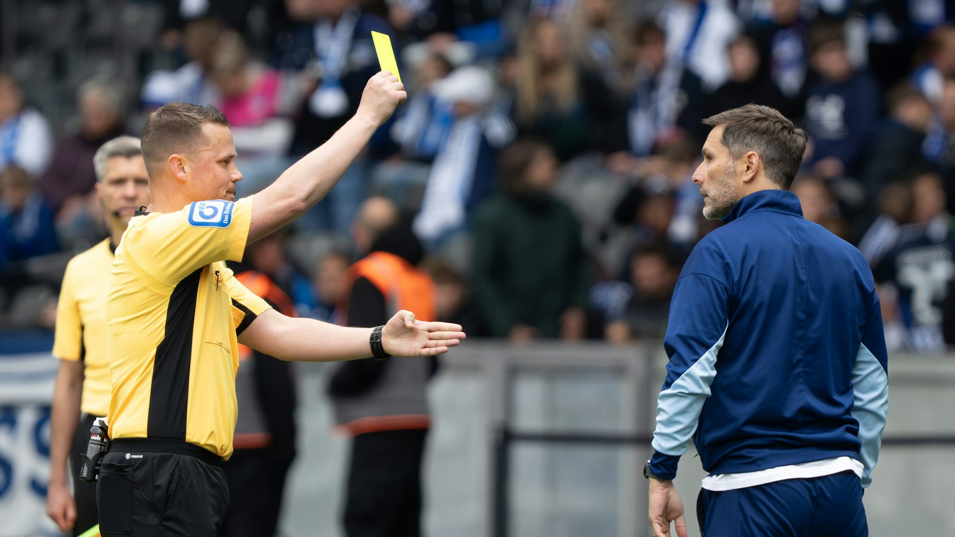 Schiedsrichter Robert Kampka (M.) zeigt Herthas Cheftrainer Stefan Leitl (r.) die gelbe Karte.
