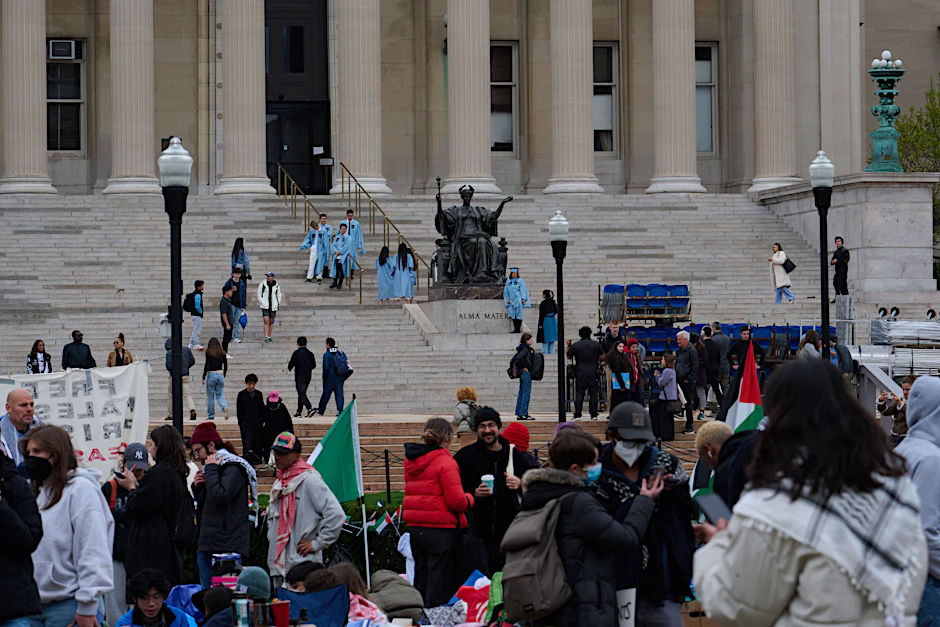 Studierende in blauen Abschlussroben posieren im April 2024 für Fotos auf den Stufen der Columbia University. Im Hintergrund halten pro-palästinensische Proteste an.