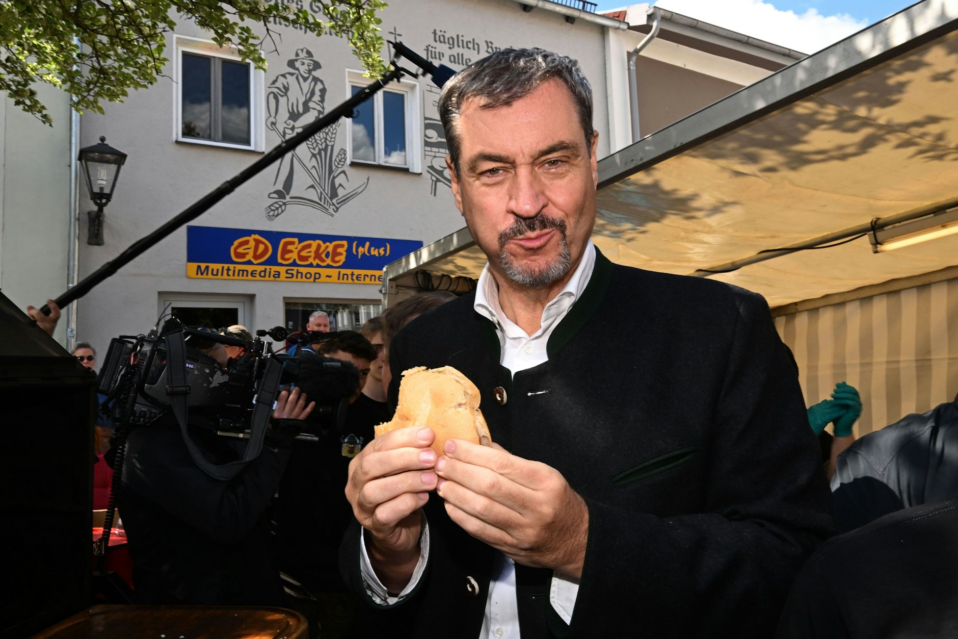 Der bayerische Ministerpräsident Markus Söder (CSU) posiert mit einer Fleischsemmel neben einem Grillmeister beim Schweine-Wettgrillen in Viechtach.