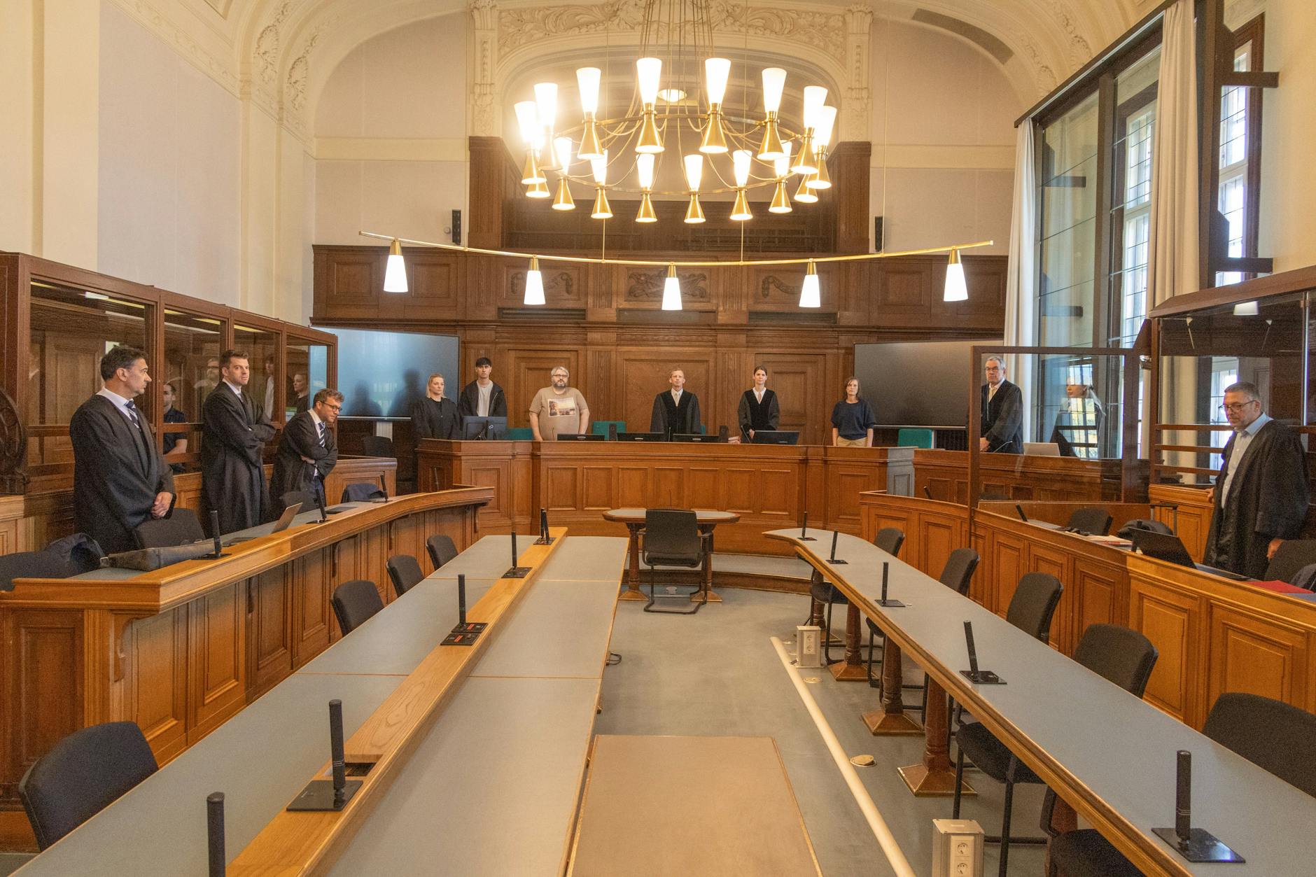 Im Gerichtssaal fehlen die Angeklagten. Rechts und links stehen die Verteidiger, in der Mitte die Richter.